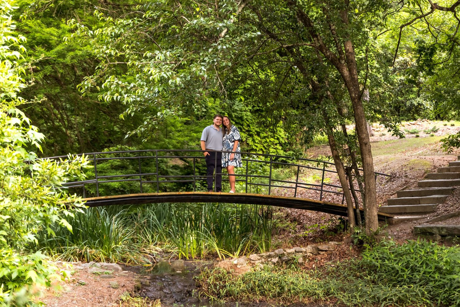 Couple standing on rustic wooden bridge surrounded by lush green forest.