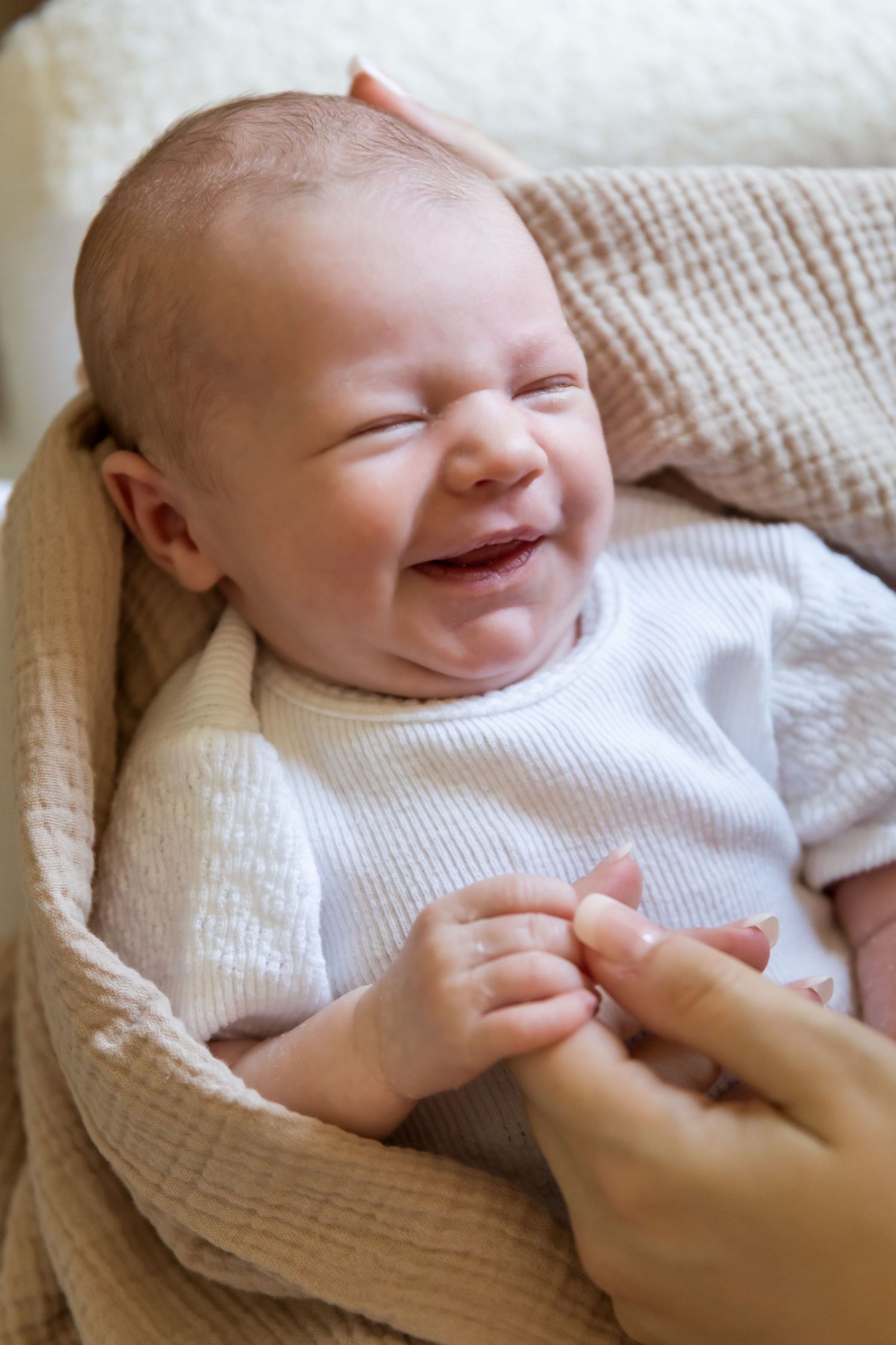 A newborn baby in a white onesie smiles while lying on a soft blanket, with an adult gently holding their tiny hands.