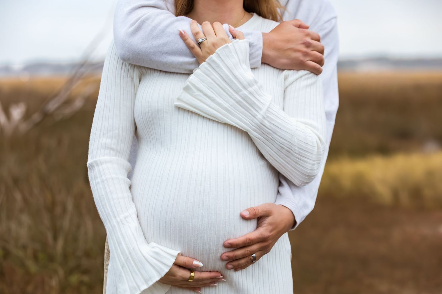 Woman in white sweater cradles baby bump while standing in golden field during sunset maternity photoshoot.