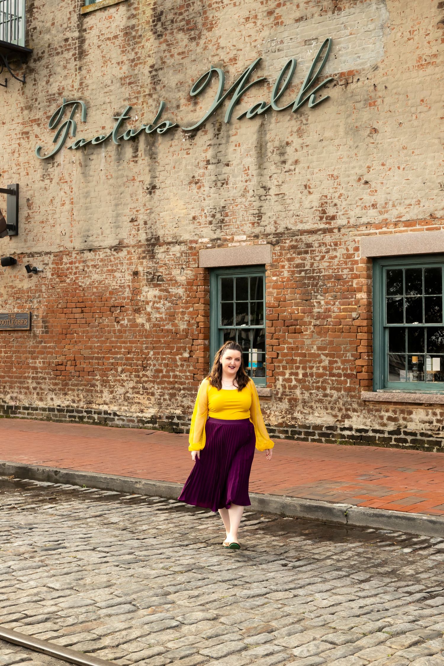 A person in a yellow top and purple skirt stands on cobblestones in front of a historic brick building with vintage signage.
