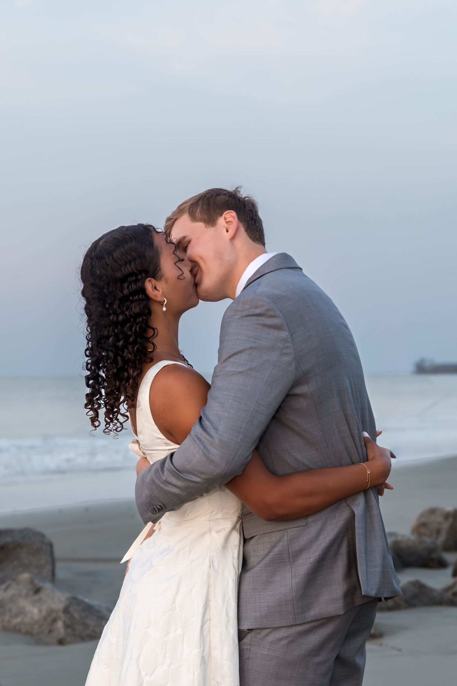 Interracial couple sharing a romantic kiss on the beach in wedding attire, with ocean waves in the background.