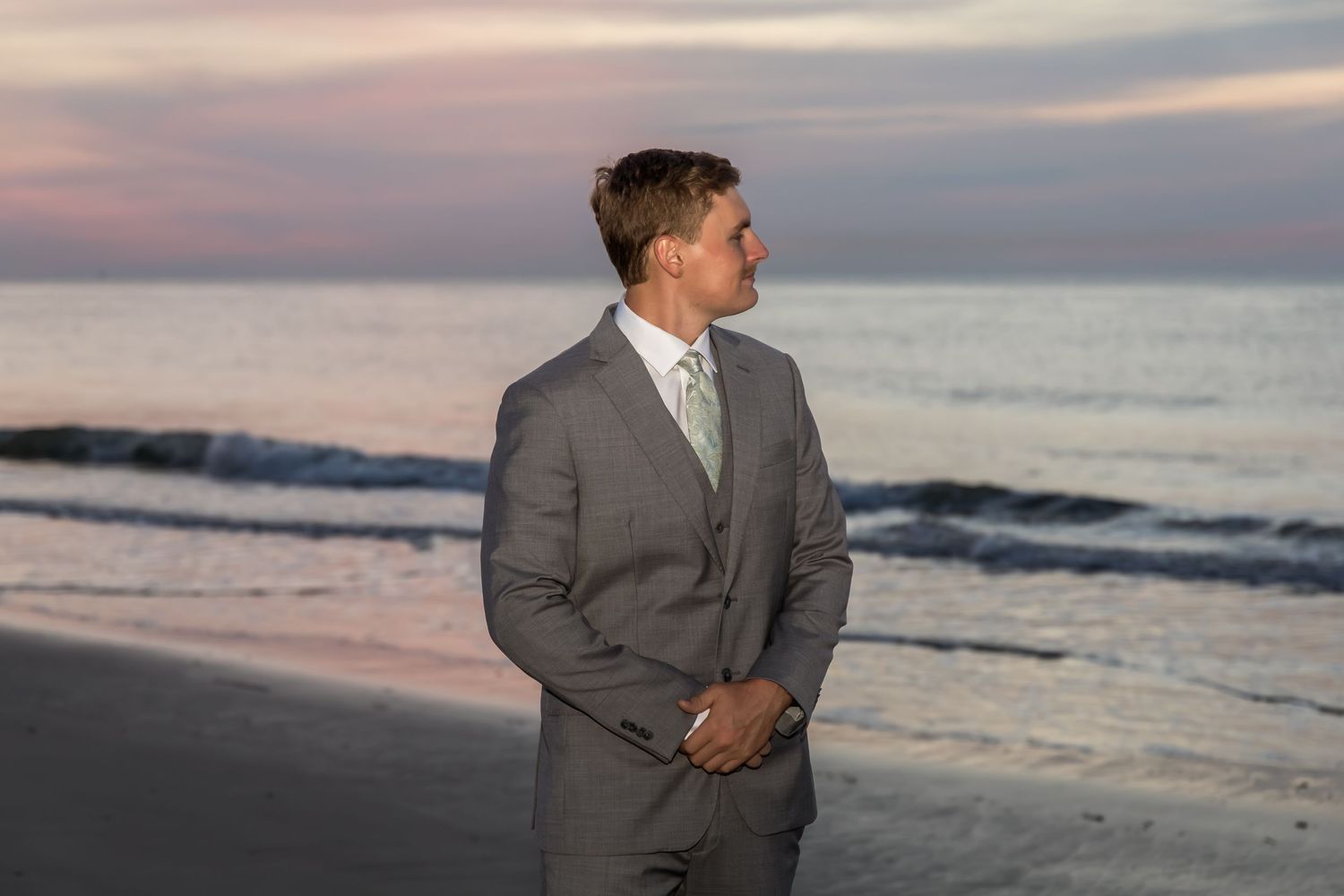 Man in gray suit standing on beach at sunset with ocean waves in background.