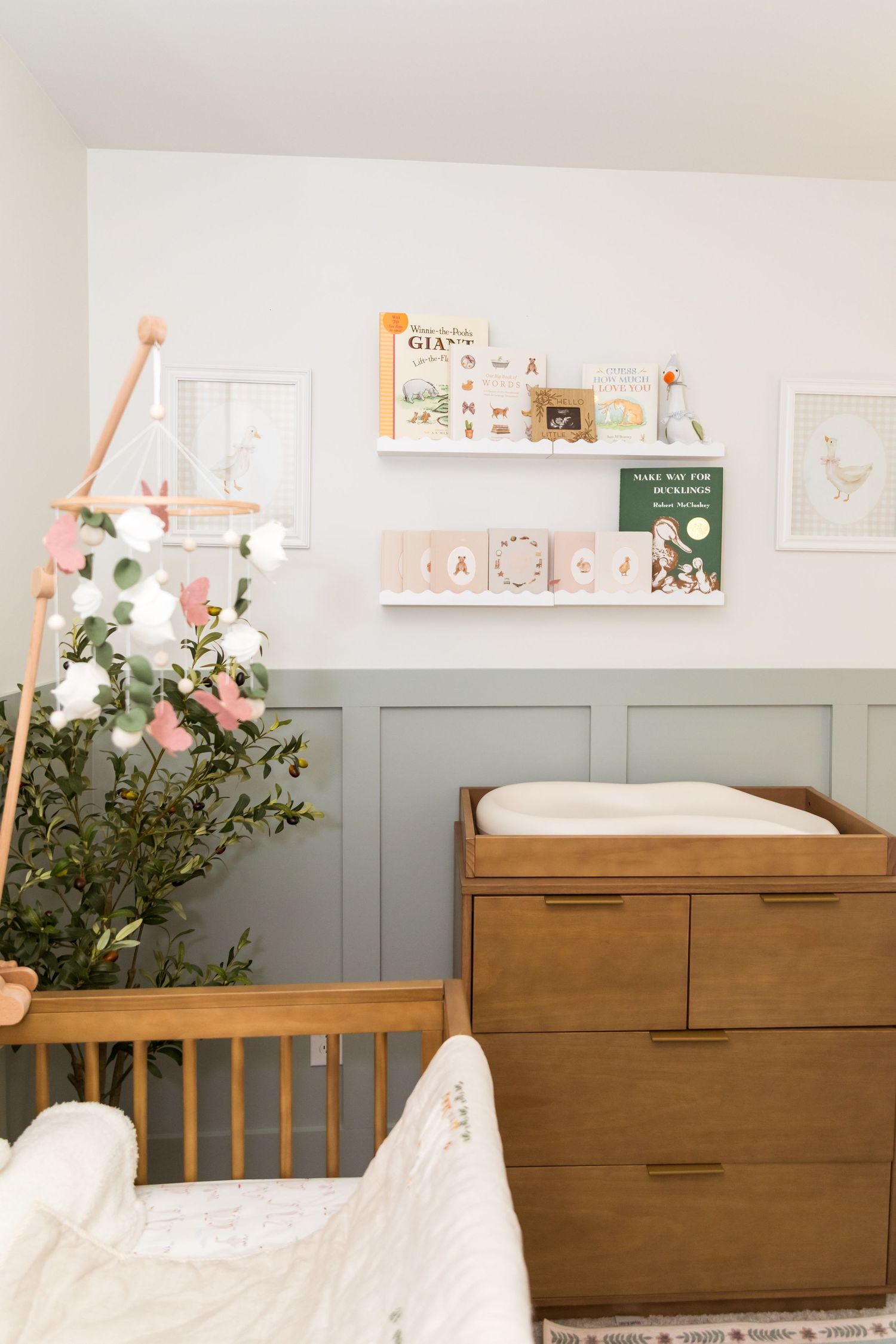 Neutral nursery room with wooden crib, dresser changing table, floating shelves with books, and floral mobile in sage green and white.