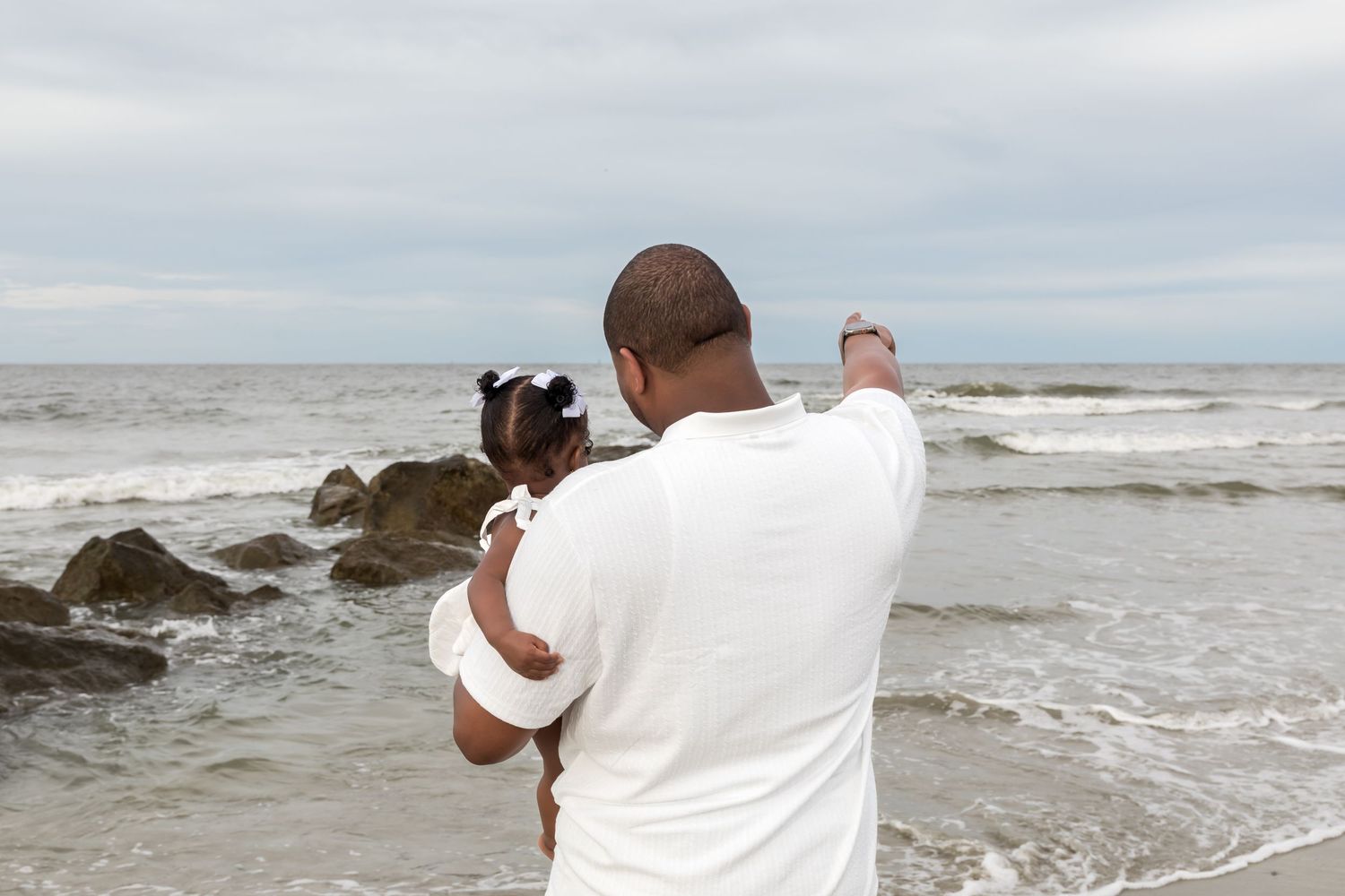 Man in white shirt holds young girl while photographing ocean waves at rocky beach on cloudy day.