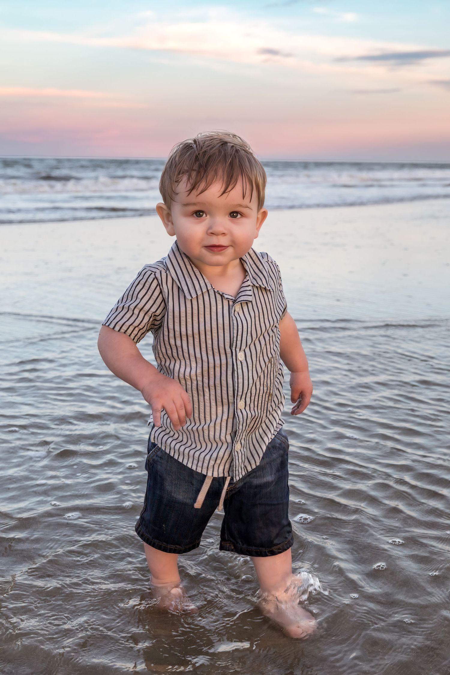 Toddler boy in striped shirt and denim shorts playing in ocean waves at sunset on a sandy beach.