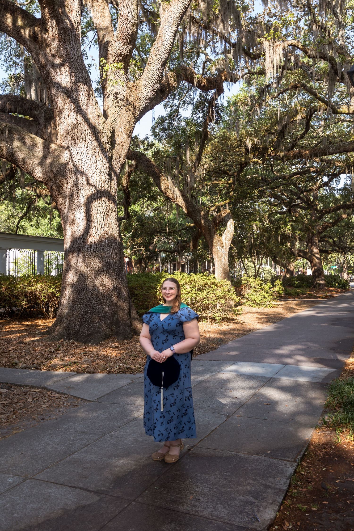 Woman in blue floral dress standing on a sidewalk beneath sprawling oak trees draped with Spanish moss.