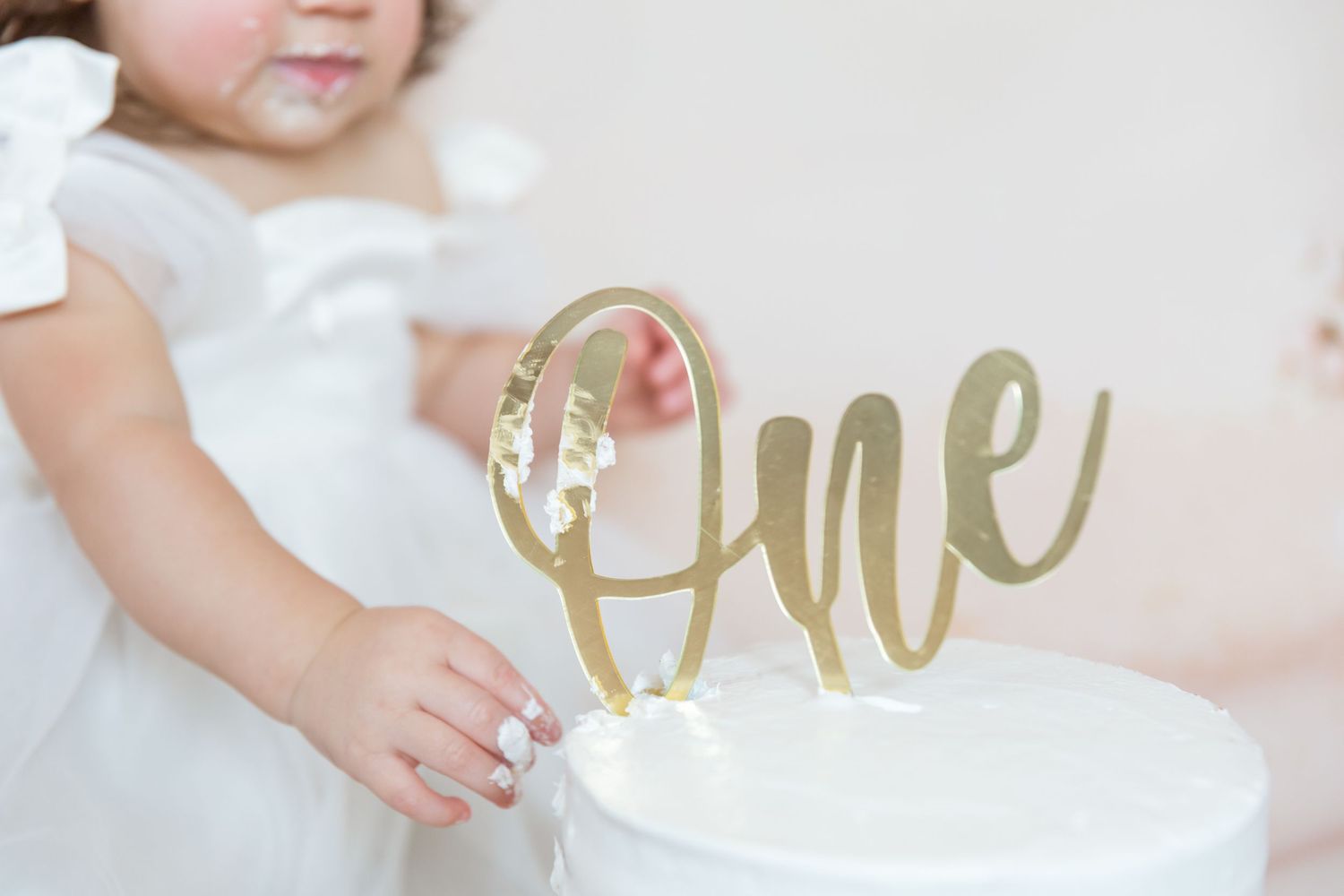 A close-up photo shows hands holding a gold 'one' cake topper decoration against a white background.
