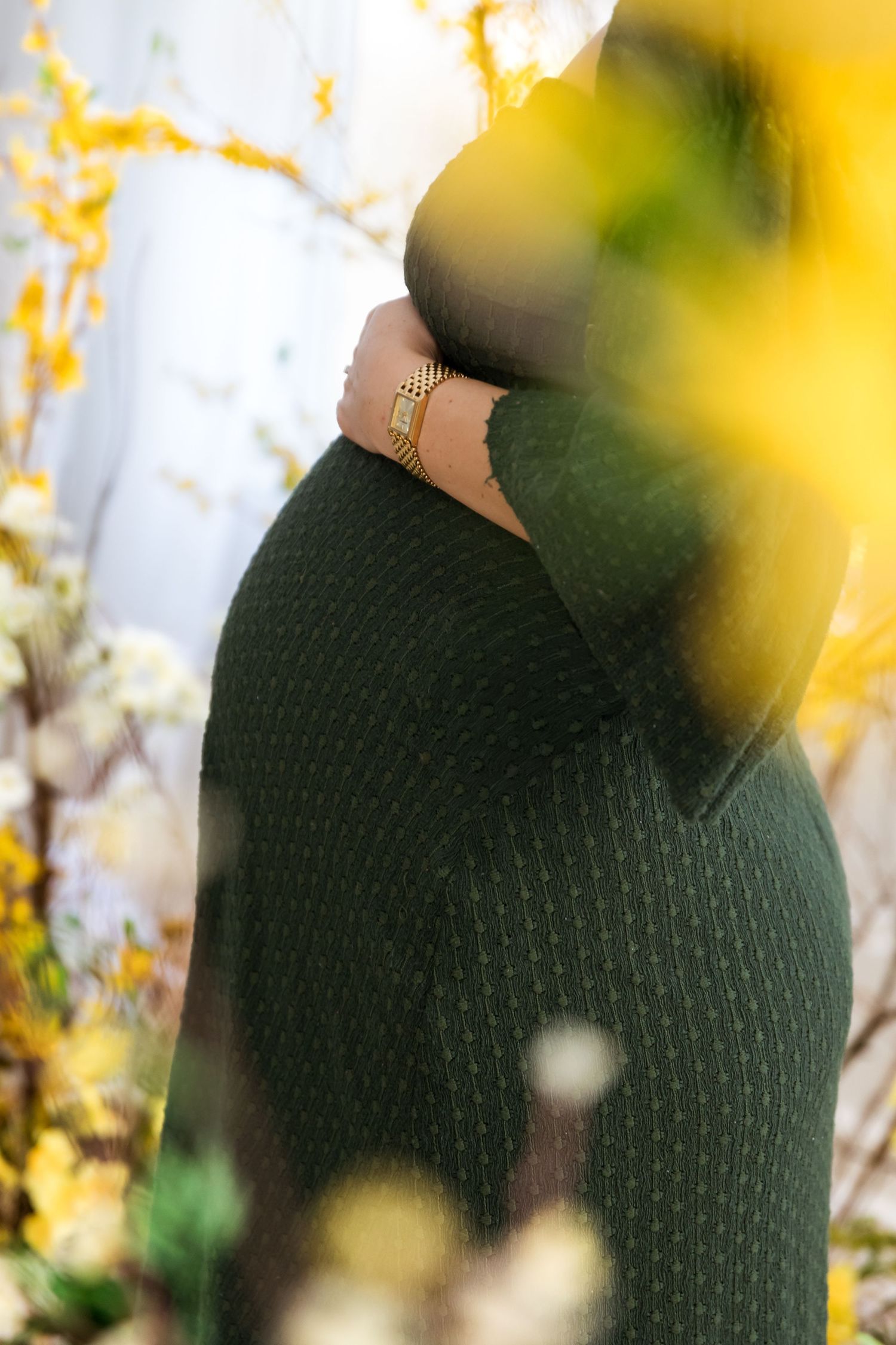 Pregnant woman in green knit sweater with gold bracelet cradling baby bump, surrounded by yellow spring flowers.