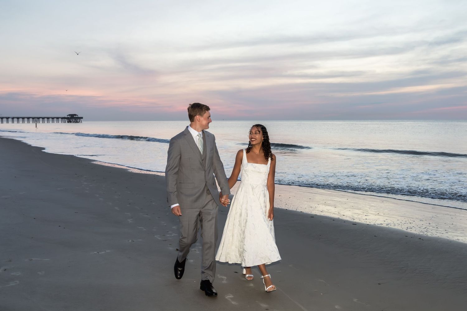 Couple holding hands walking on beach at sunset, man in gray suit and woman in white dress near ocean pier.