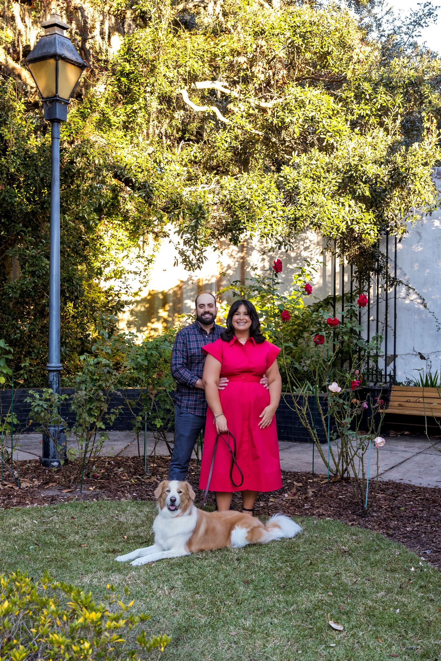 A couple and their golden retriever dog pose together on a grassy lawn in front of bushes and a garden setting.
