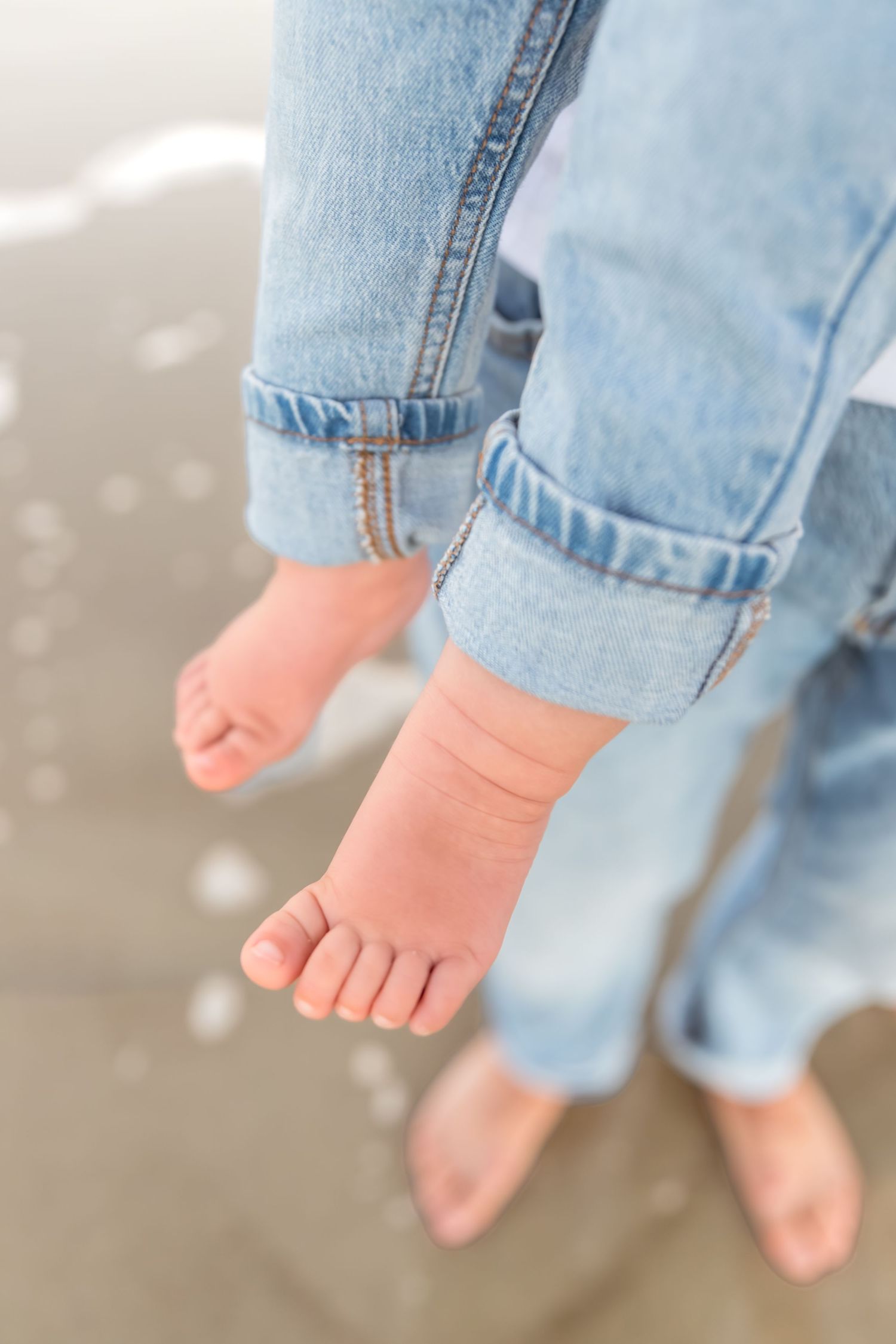 Small bare feet of a child wearing rolled up light blue jeans while standing on a wet surface.