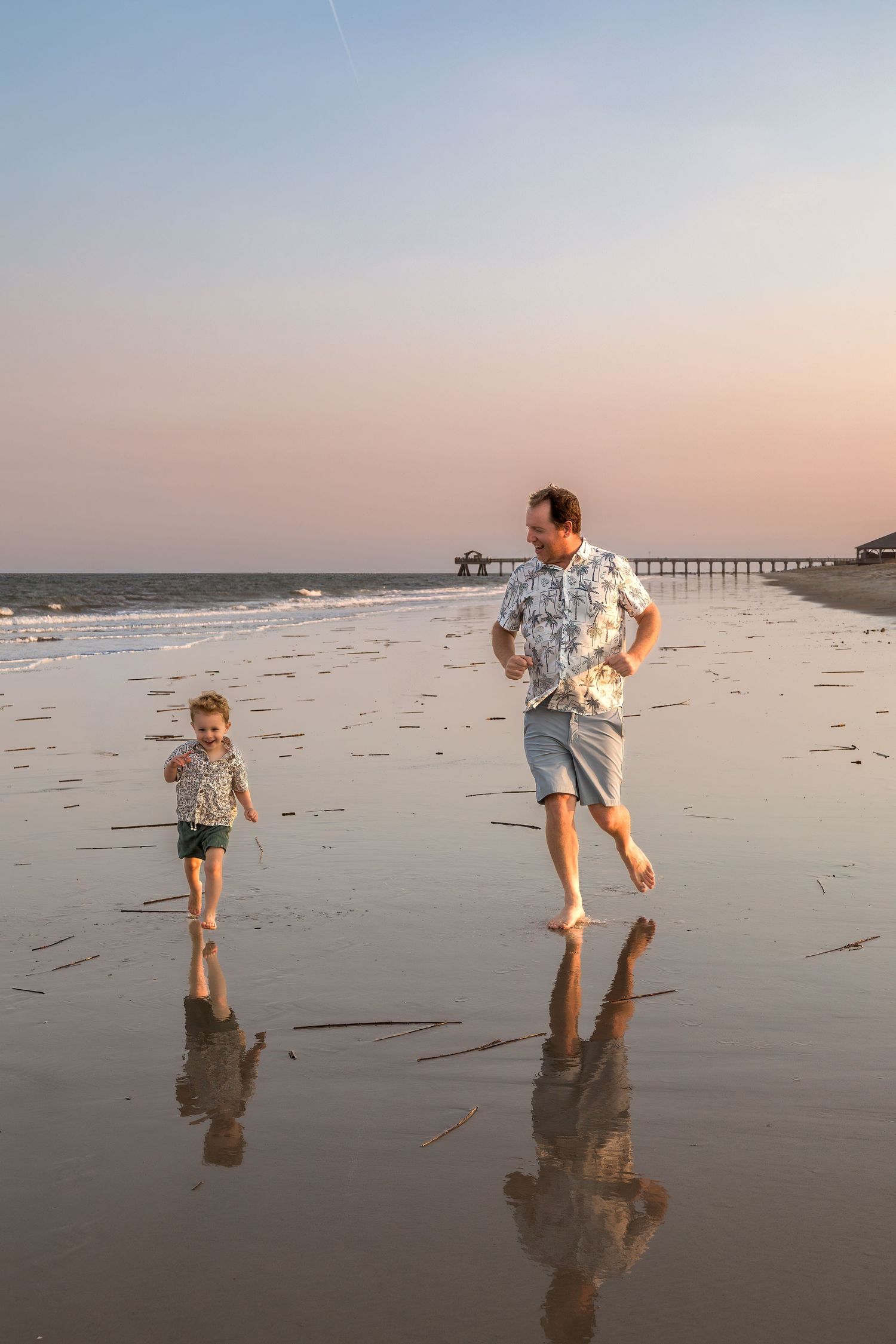 Father and child running barefoot on a wet sandy beach at sunset with a pier visible in the background.