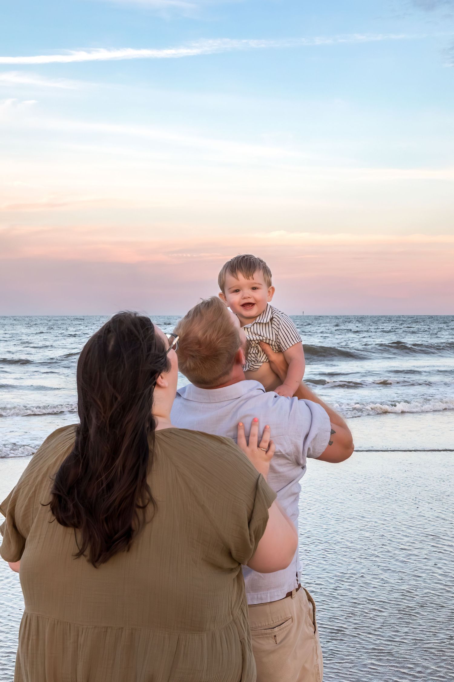A couple stands on a beach at sunset, holding their smiling baby up in the air near the ocean waves.