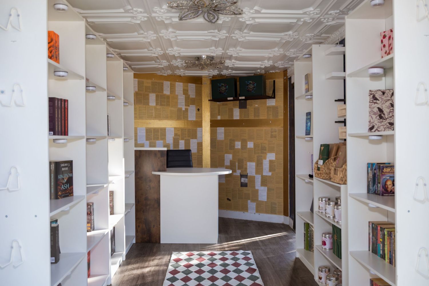 Cozy bookstore interior with white shelves, golden mosaic accent wall, checkout desk, and checkered floor rug.