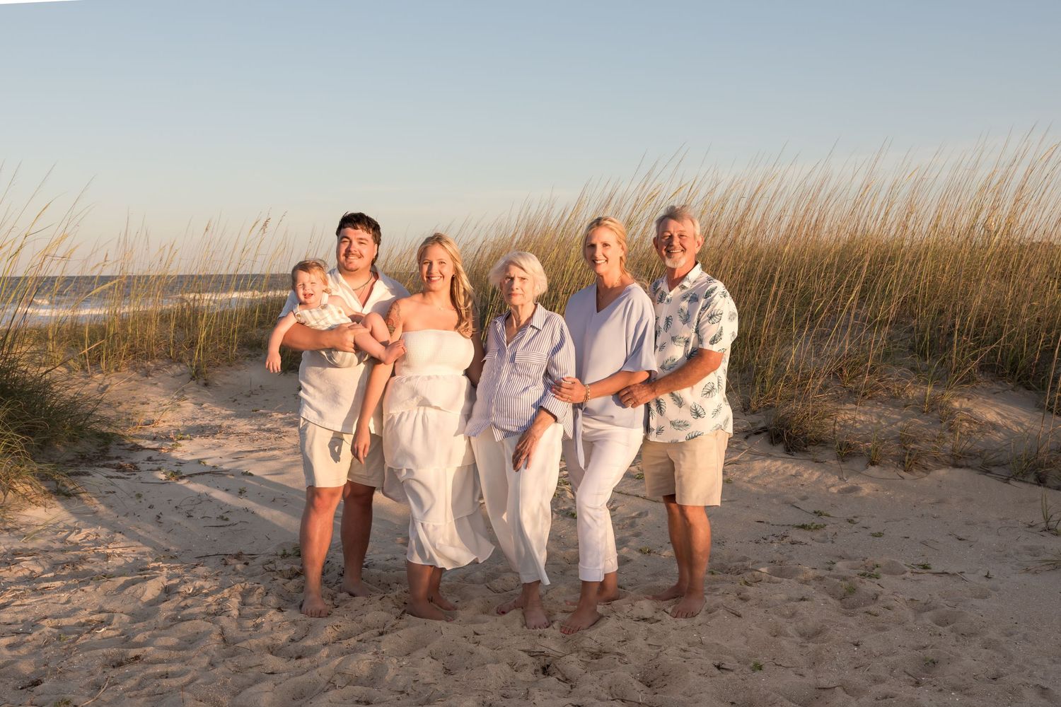 Multi-generational family smiling together on a sandy beach at sunset with tall grass in the background.