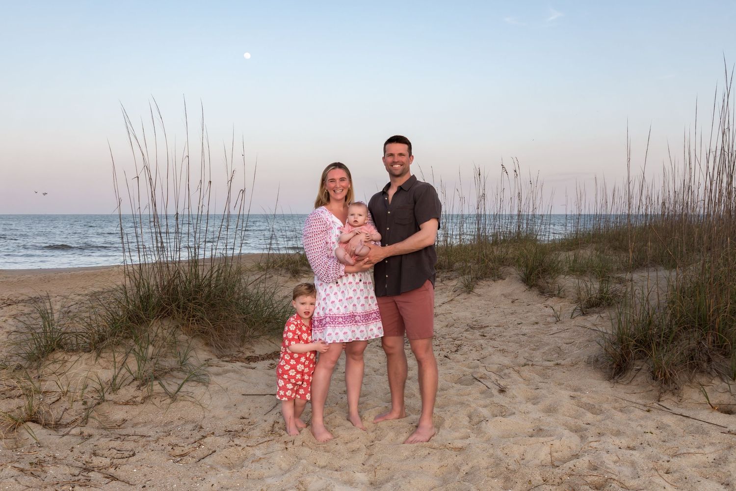Family of four poses on sandy beach at sunset with sea oats and ocean in background.