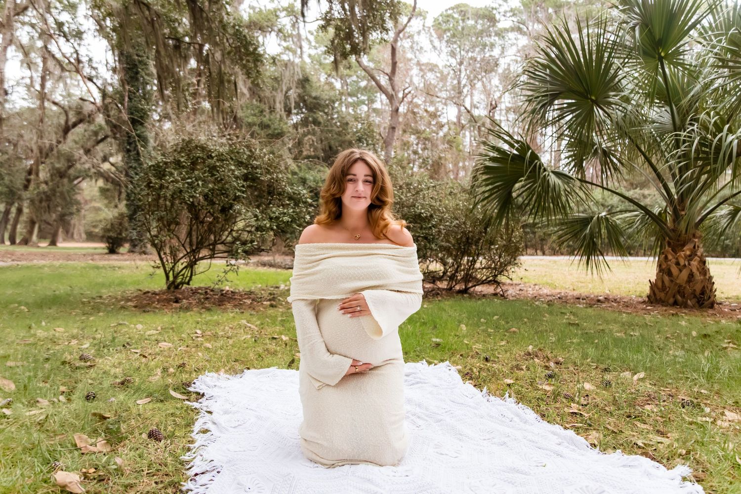A person in a white off-shoulder wedding dress poses gracefully in a garden setting with palm trees and greenery.