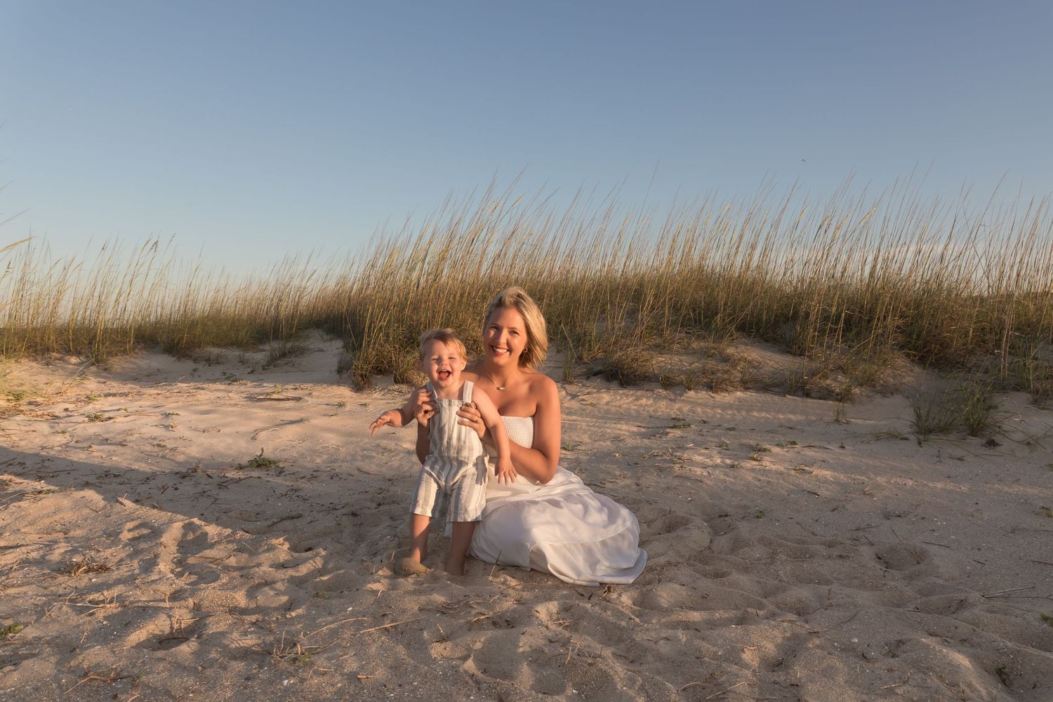 A smiling woman in a white dress sits on sandy beach dunes with a happy baby beside her at golden hour.