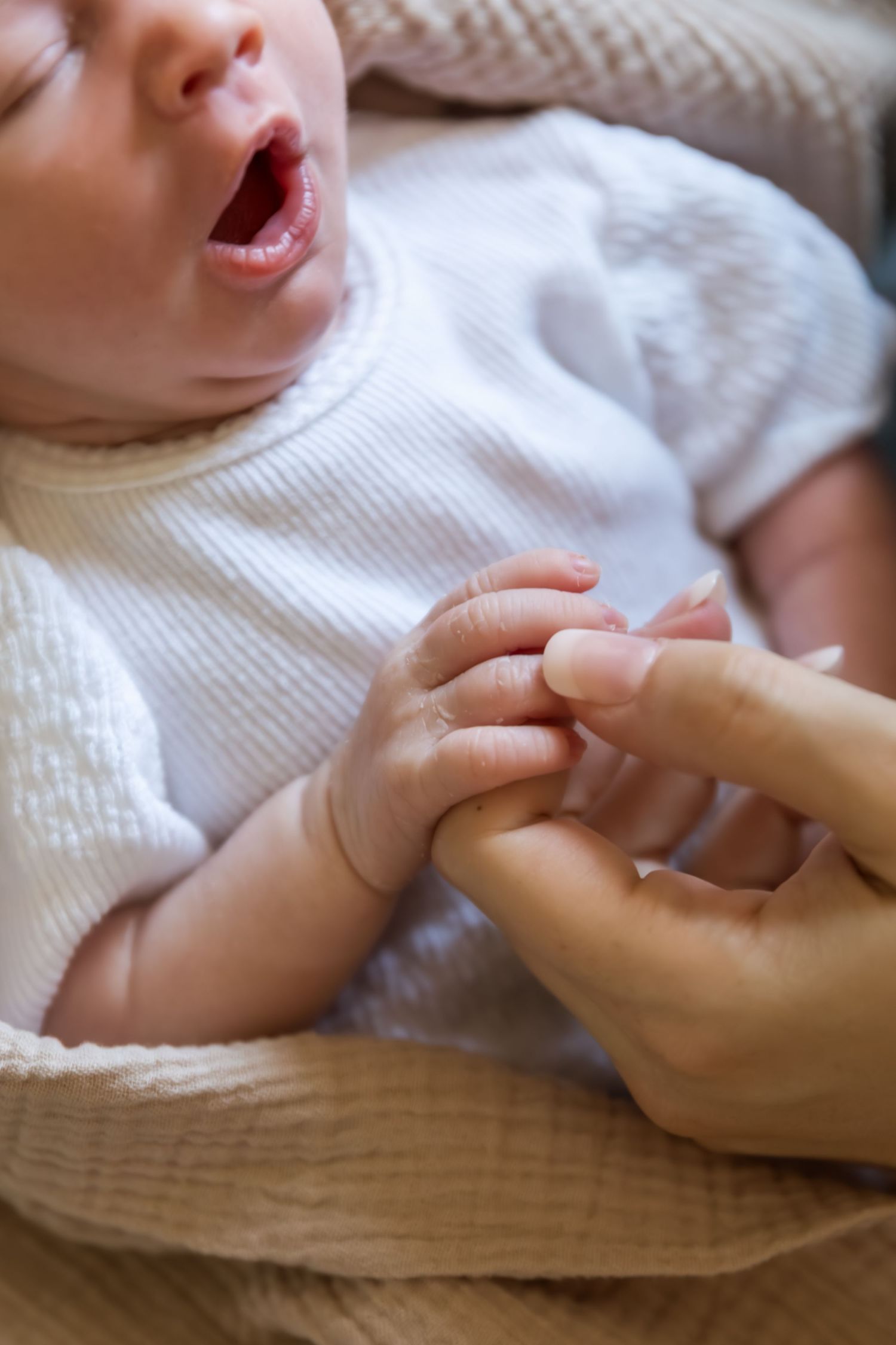 Newborn baby in white onesie crying while holding an adult finger, close-up of tiny hand gripping parent.