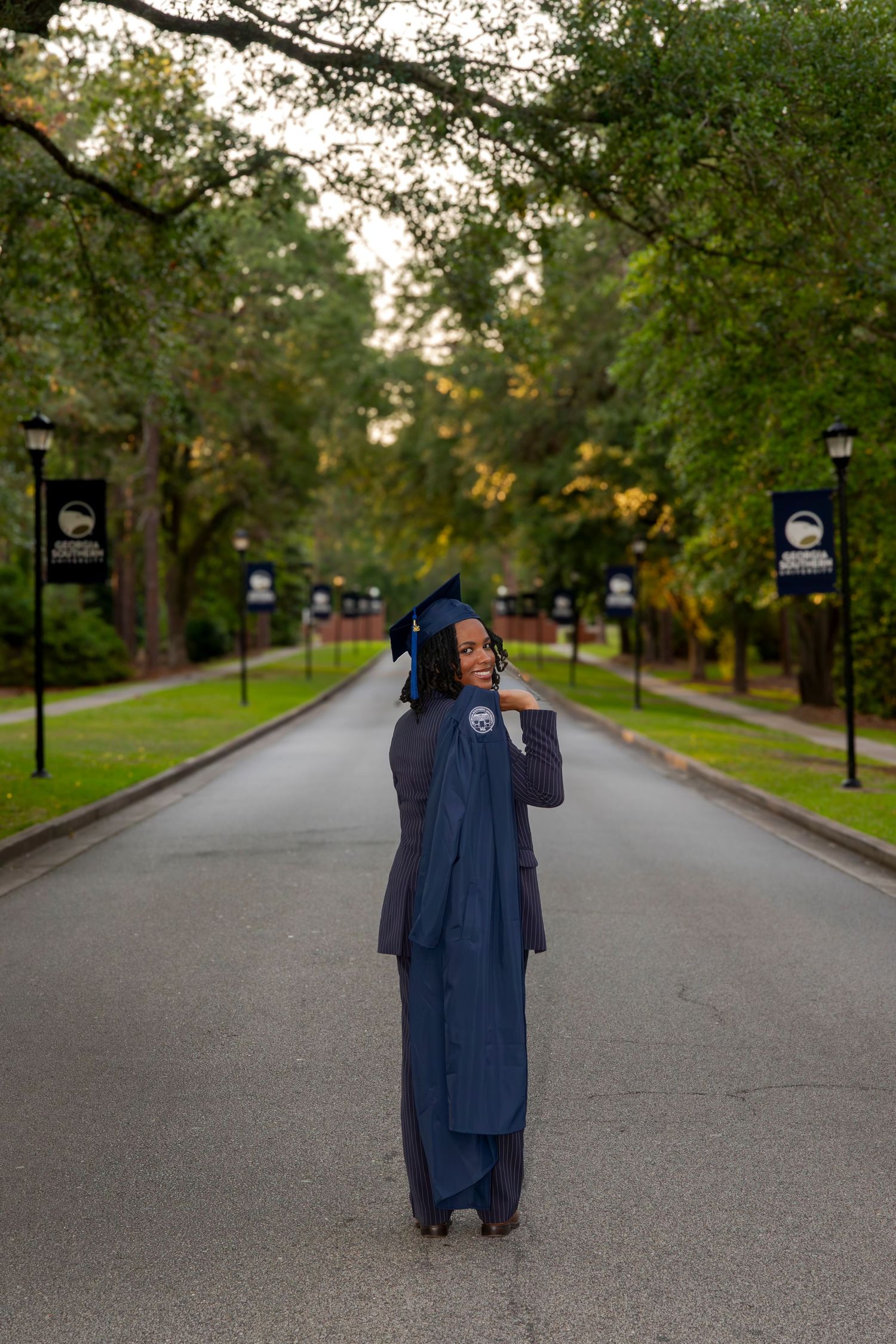 Graduate in navy blue cap and gown poses on tree-lined campus pathway.