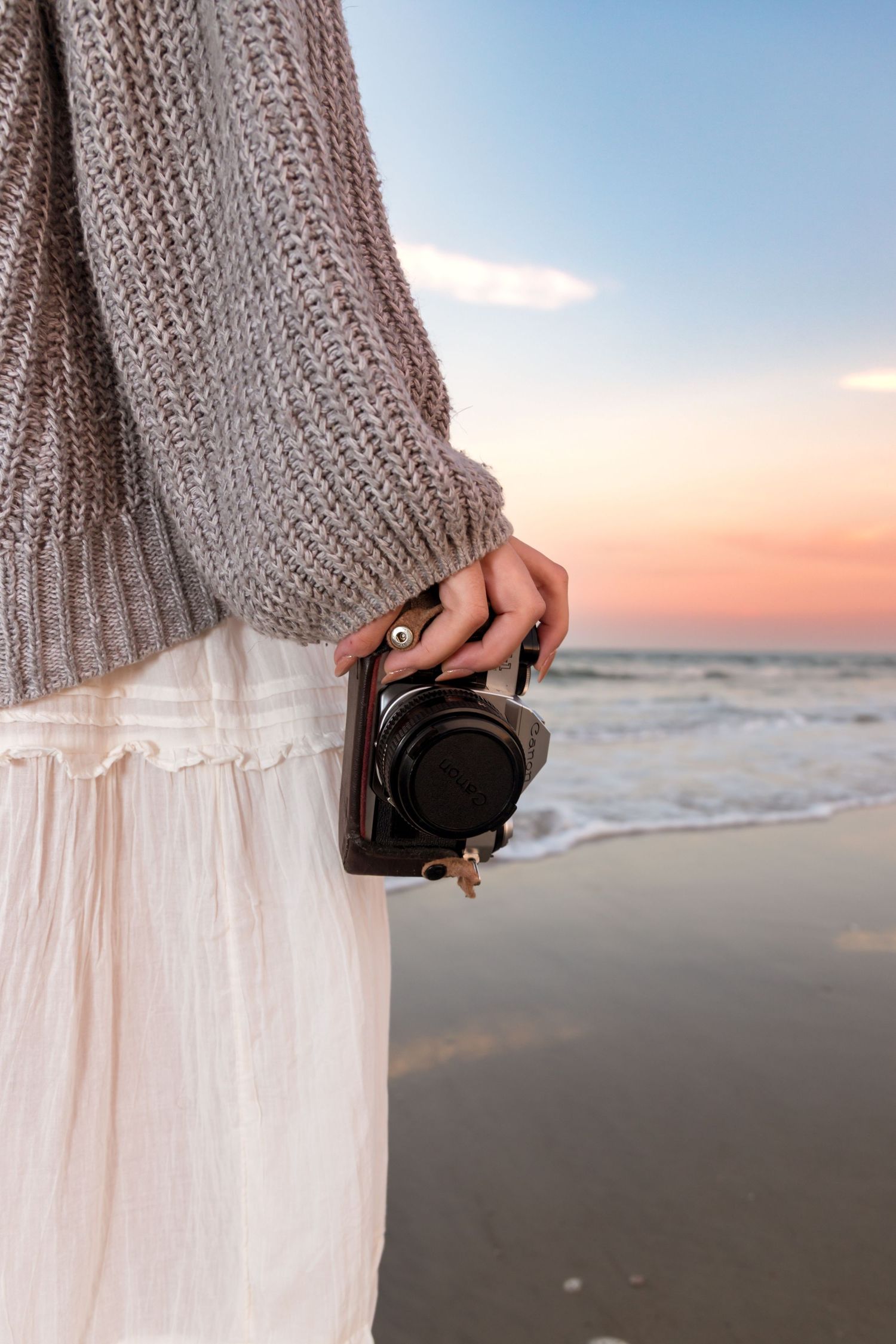Person wearing cozy sweater and flowy dress holding wallet on beach at sunset.