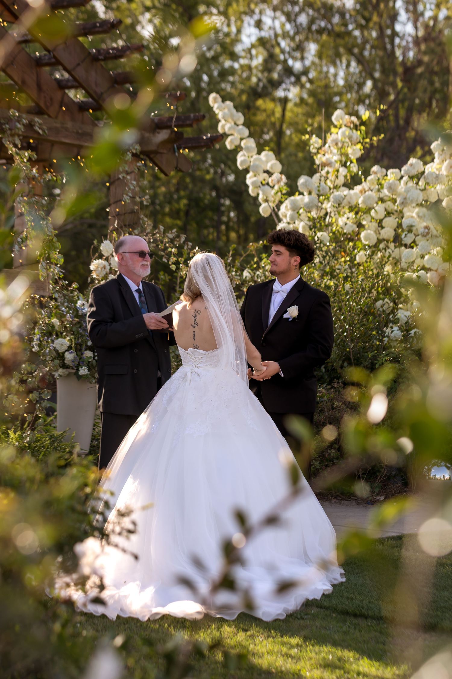 Bride and groom exchange vows outdoors surrounded by white roses and lush greenery in a garden wedding ceremony.