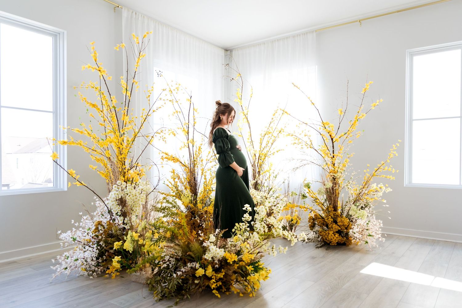 A pregnant woman in a green dress stands among lush yellow and white floral arrangements in a bright white studio.