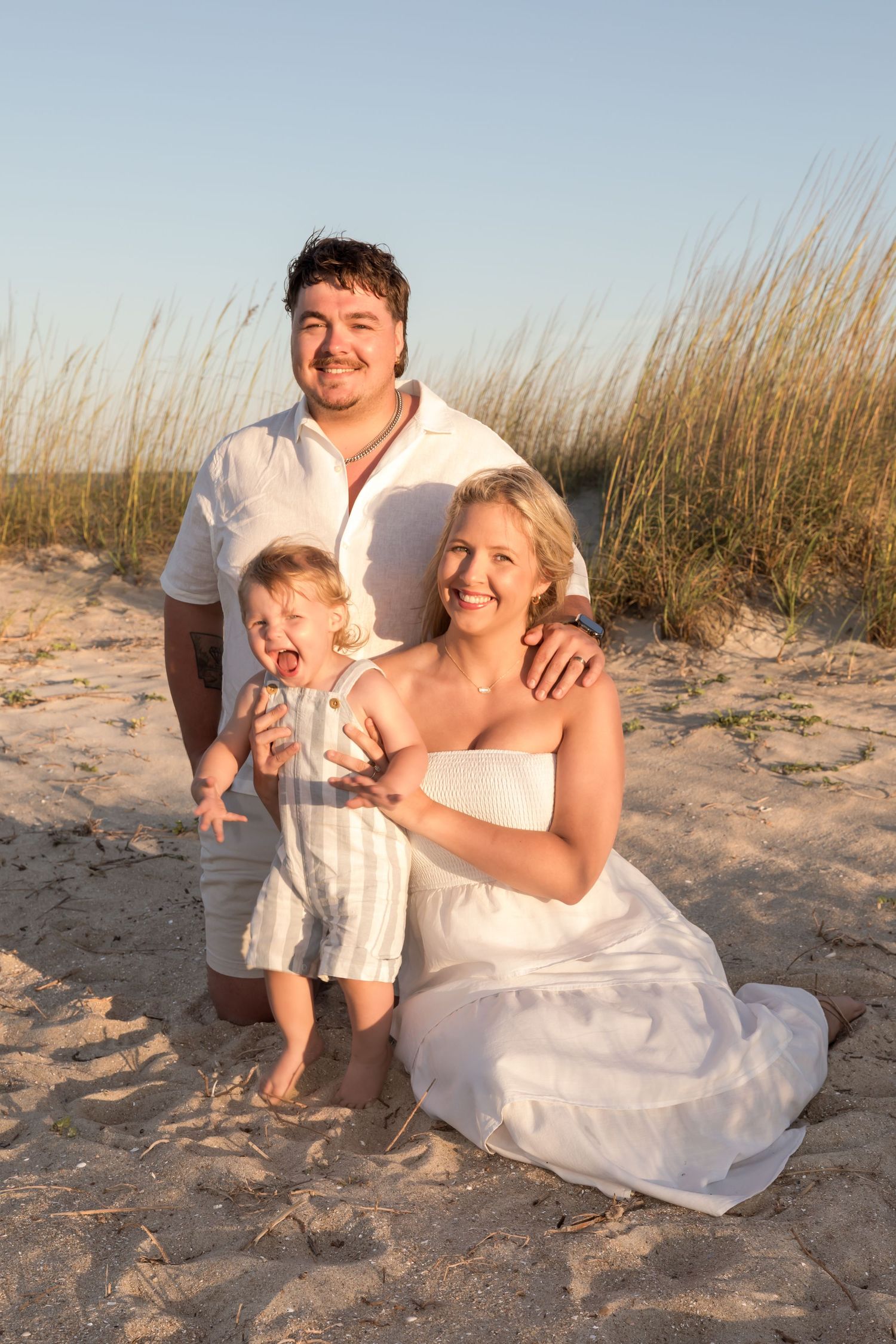 Young family with baby posing on sandy beach at golden hour near sea grass dunes.