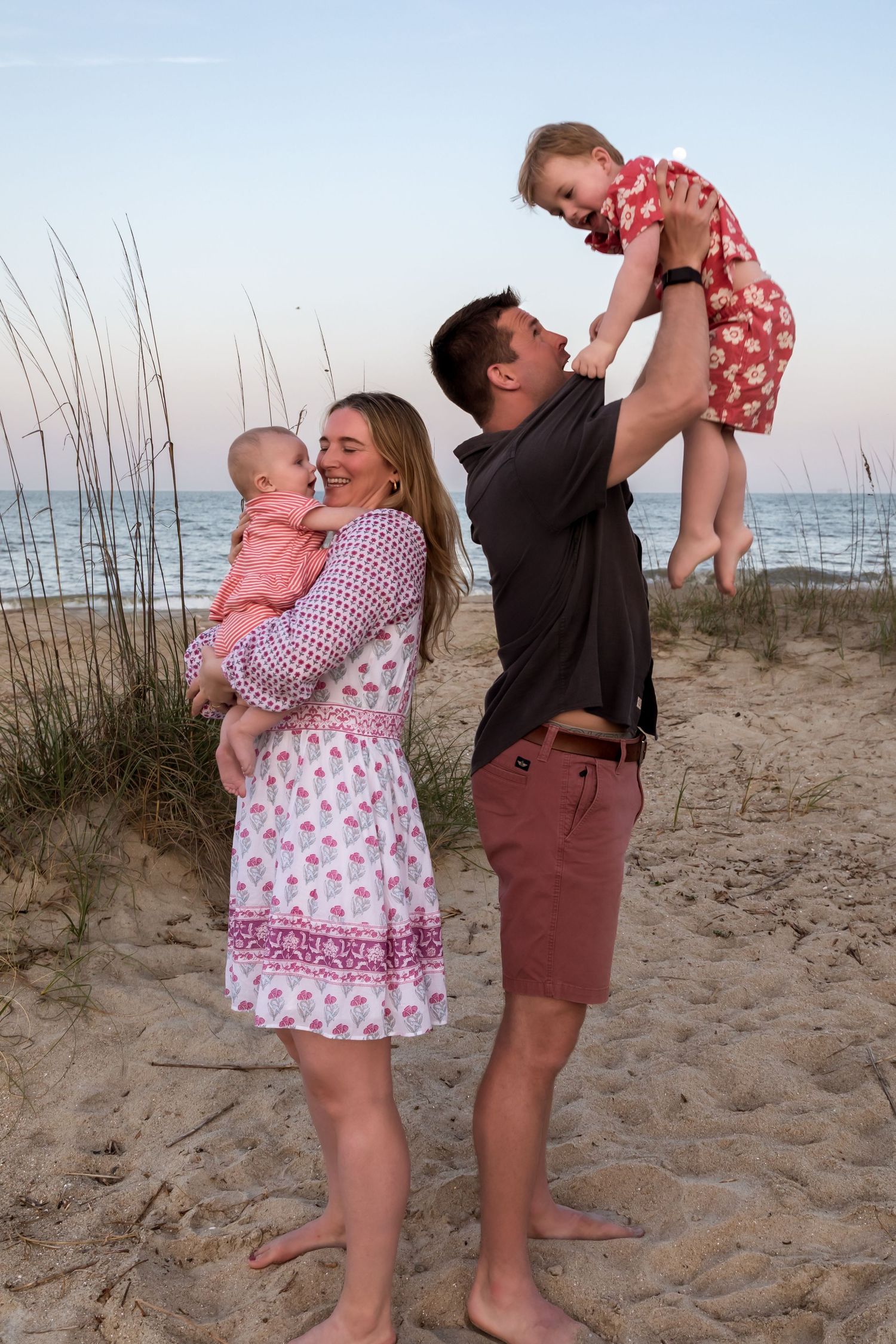 Family of four enjoying a beach sunset, parents holding two young children on sandy shore with ocean waves in background.