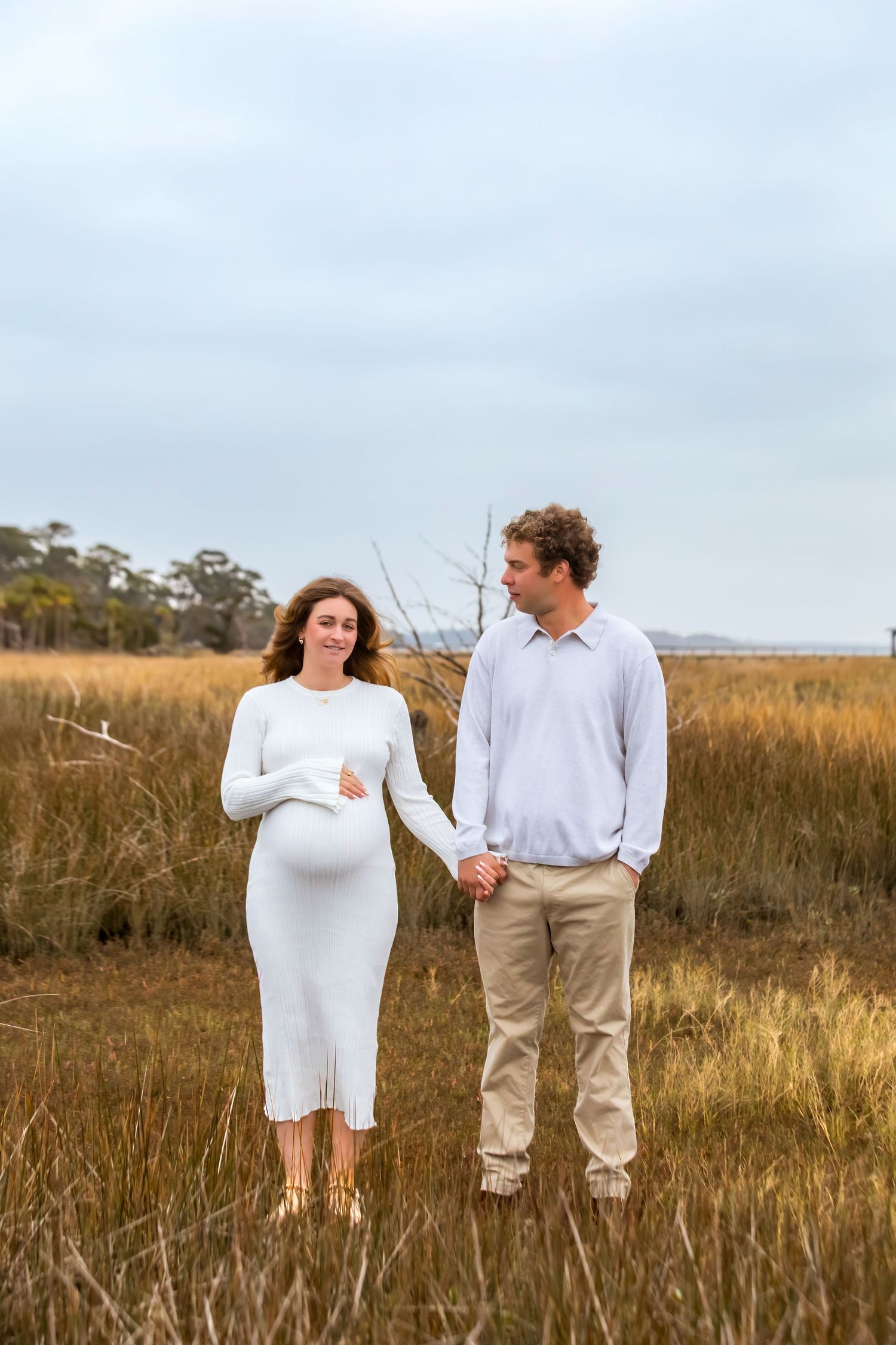 A couple in casual white and khaki attire stands together in a golden grass field during sunset.