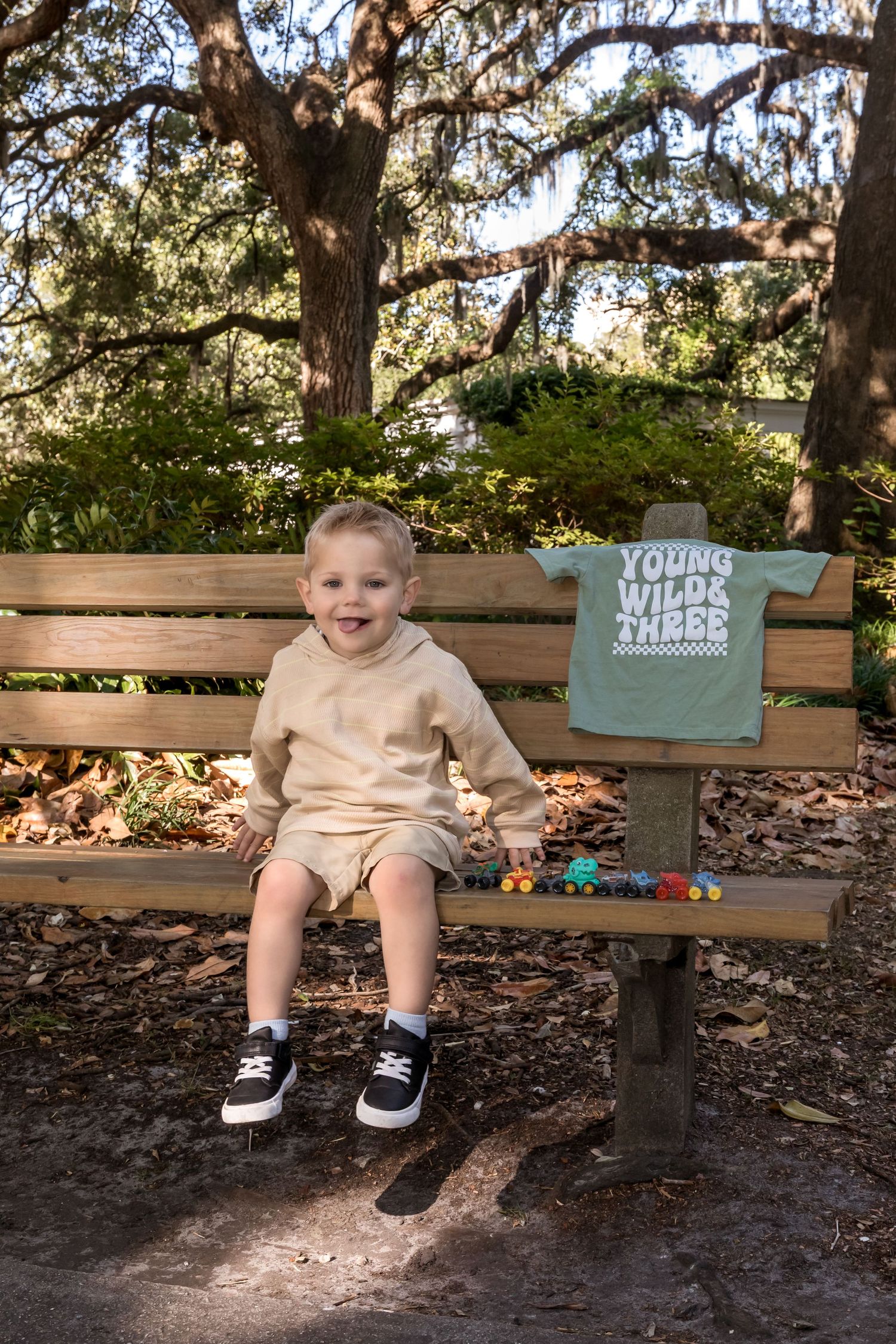Toddler boy sitting on park bench beside a Young Wild & Three shirt display, surrounded by trees.