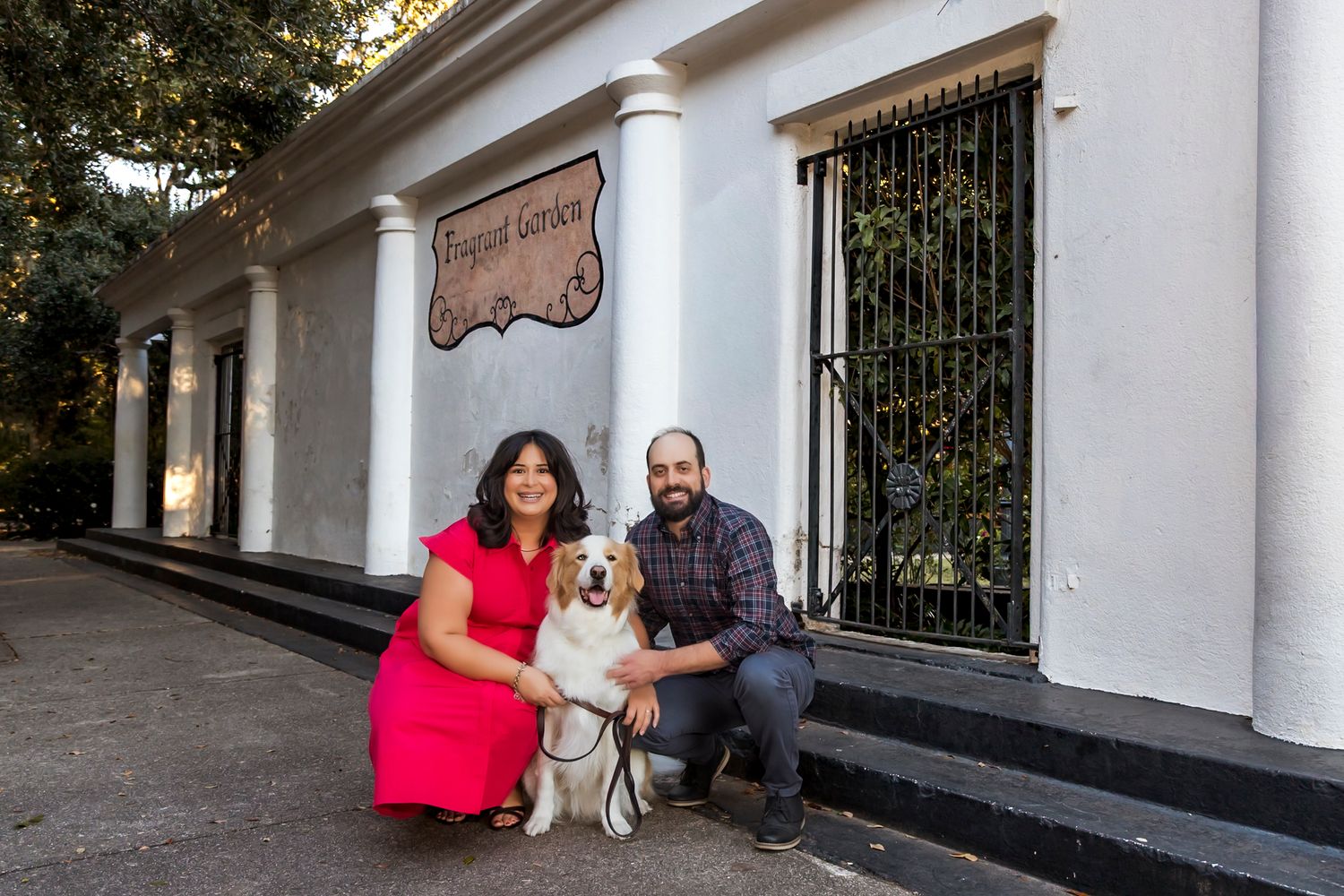 Couple with their dog posing in front of white building with wrought iron gates.