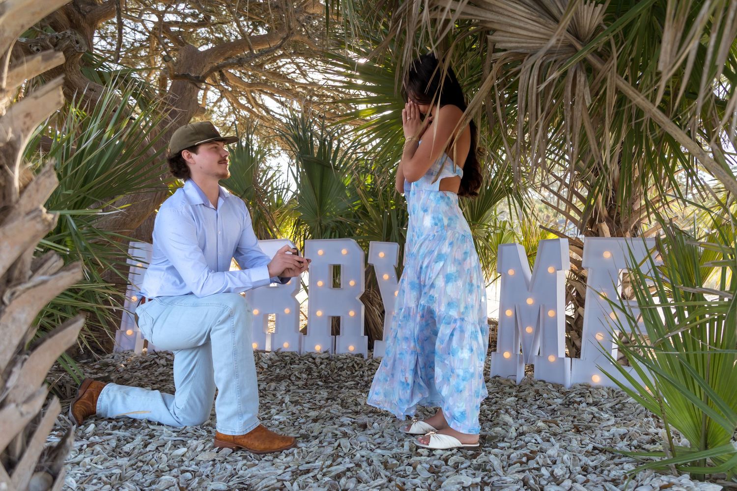 Man kneeling proposing to woman in blue floral dress near lit marquee letters outdoors among palm trees.