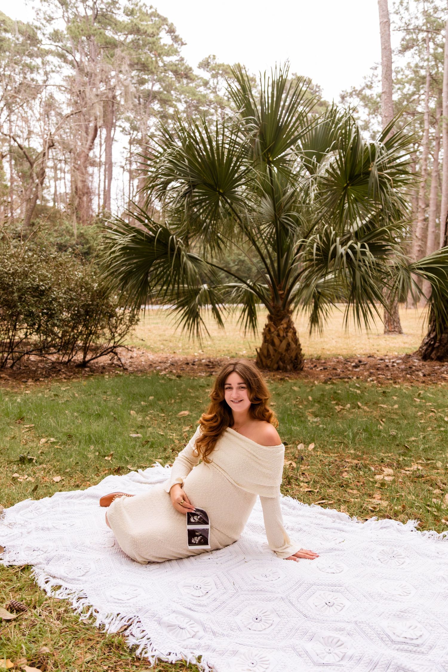 A white lace dress flows across a blanket on grass with palm trees and pine trees in the background.