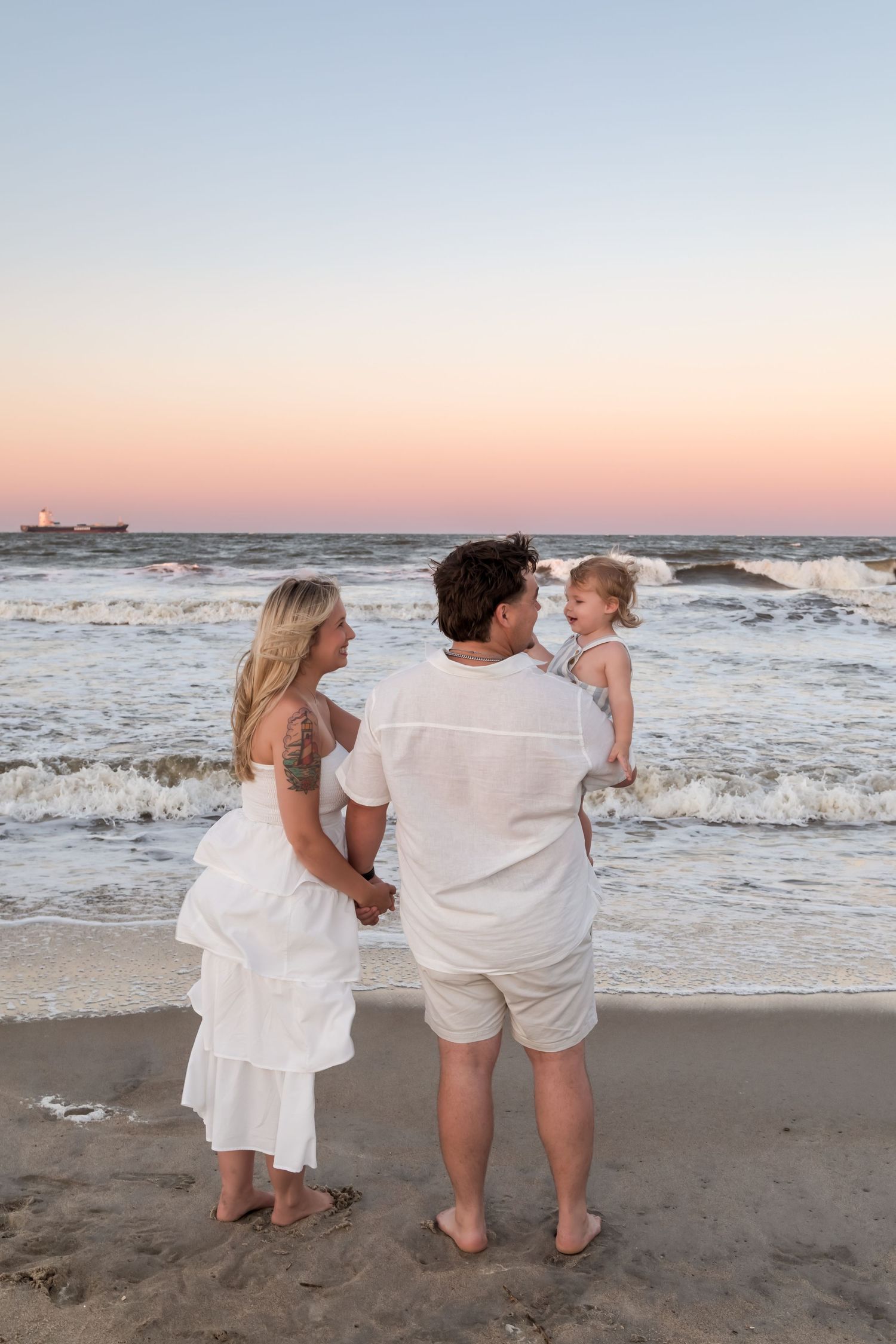 Family of three in white outfits standing on a beach at sunset, parents holding toddler by ocean waves.