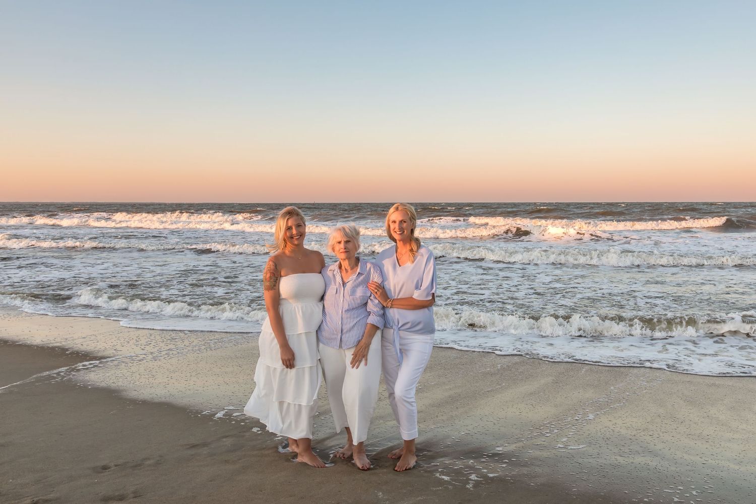Three women smiling together on a sandy beach at sunset with ocean waves in the background.