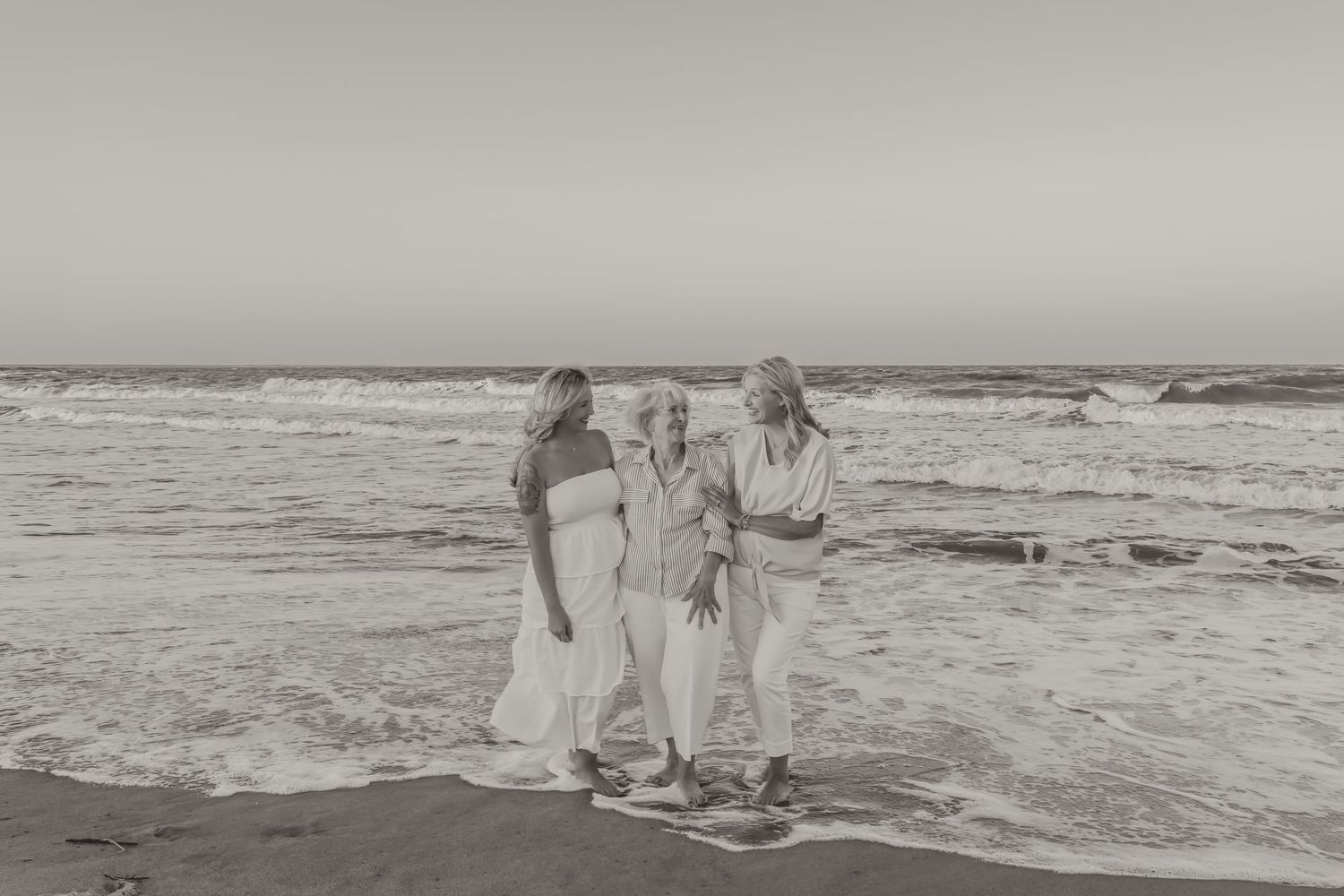 Three women embrace and walk together along a sandy beach with ocean waves in black and white photo.