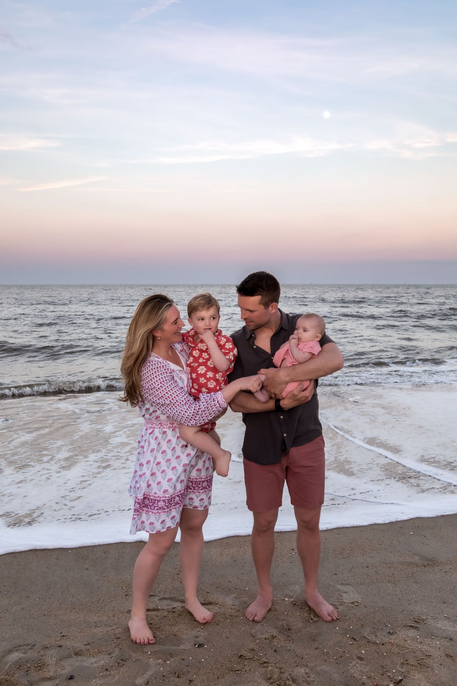Family of four poses barefoot on sandy beach at sunset, parents holding two young children with ocean waves behind them.