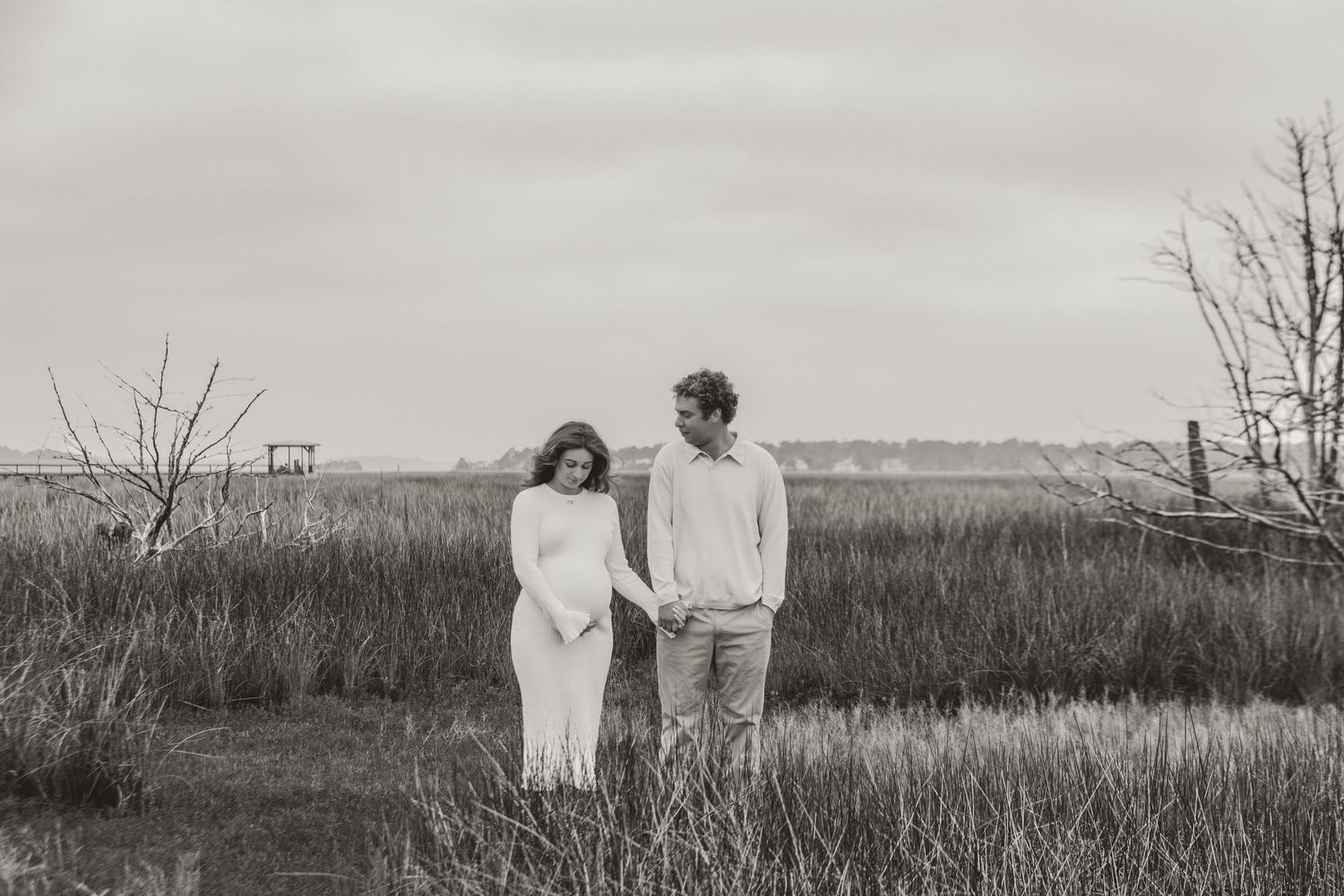 A black and white photo of a couple standing together in a rustic field with bare trees in the background.