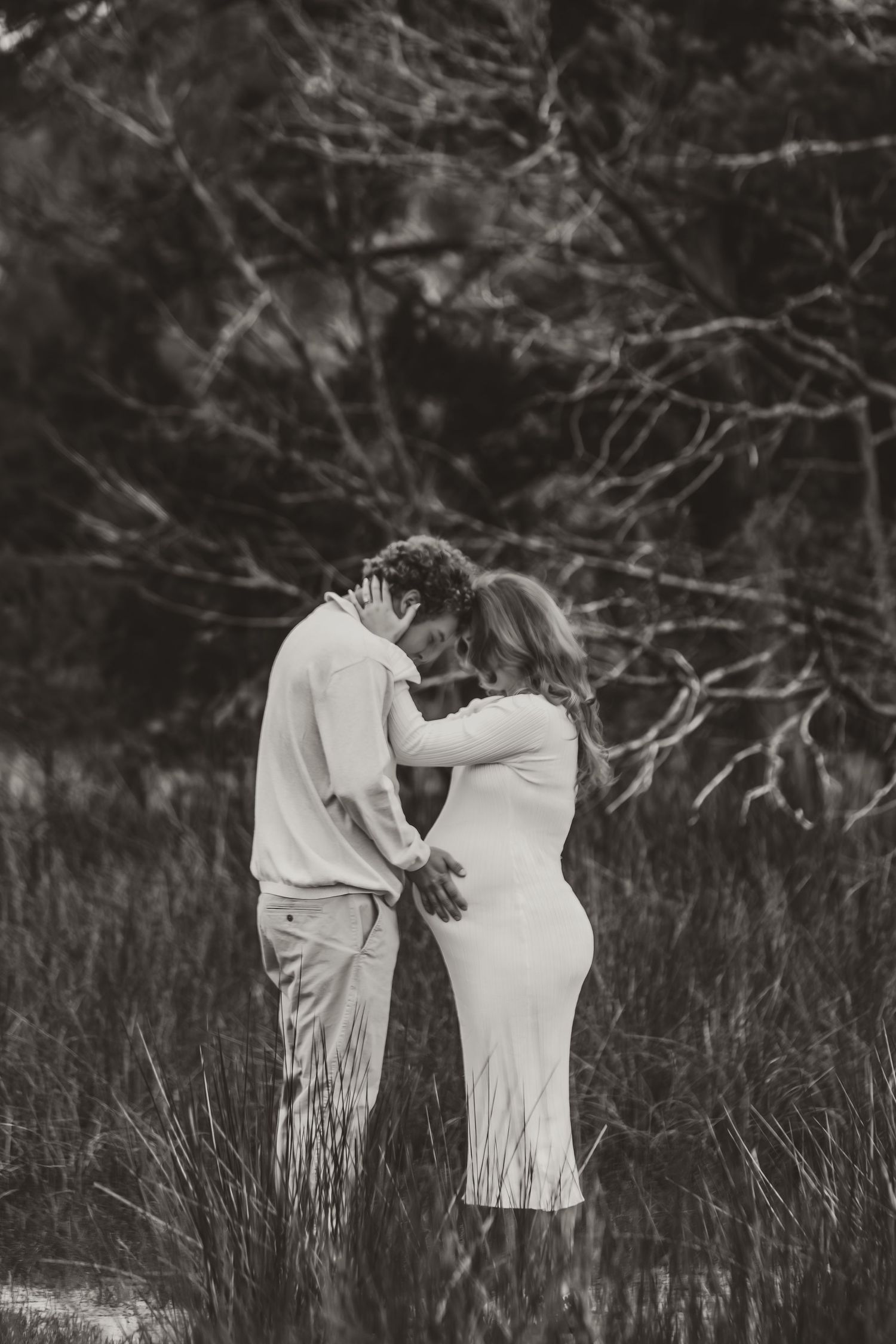 Black and white photo series of a couple in white dresses embracing in a field with bare branches in background.