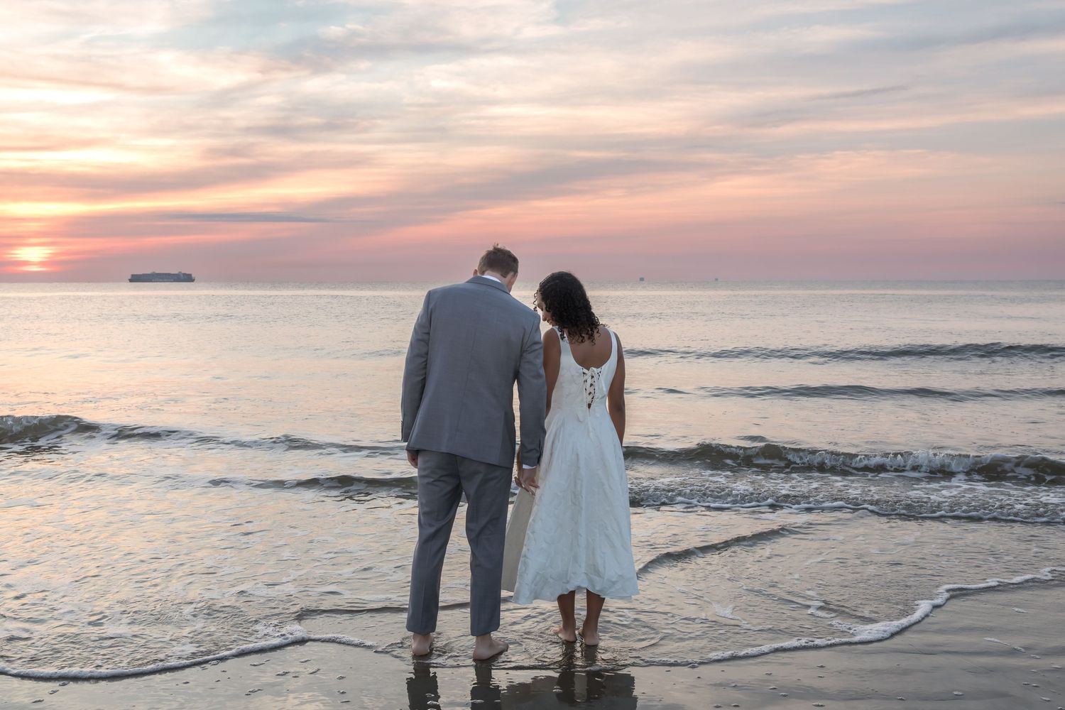 Couple holding hands on a sandy beach at sunset, woman in white dress, man in gray suit, ocean waves in background.