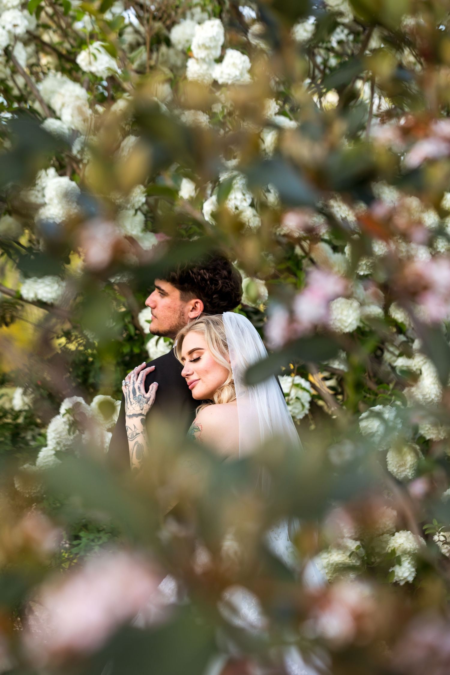 A bride and groom embrace among blooming white spring flowers, captured through lush green foliage in a romantic outdoor setting.