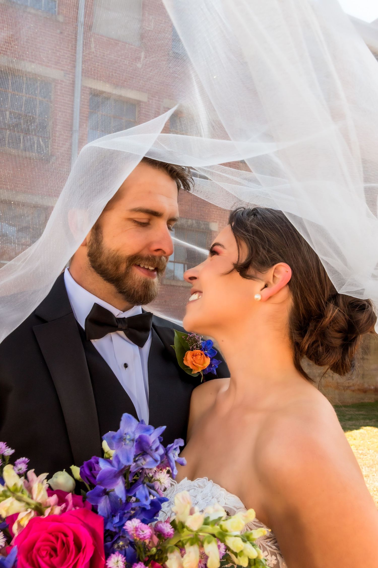 Couple embraces under a white veil with colorful wedding bouquet.