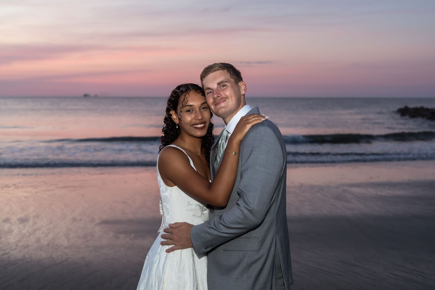 Newlywed couple embracing on a beach at sunset, bride in white dress and groom in gray suit.