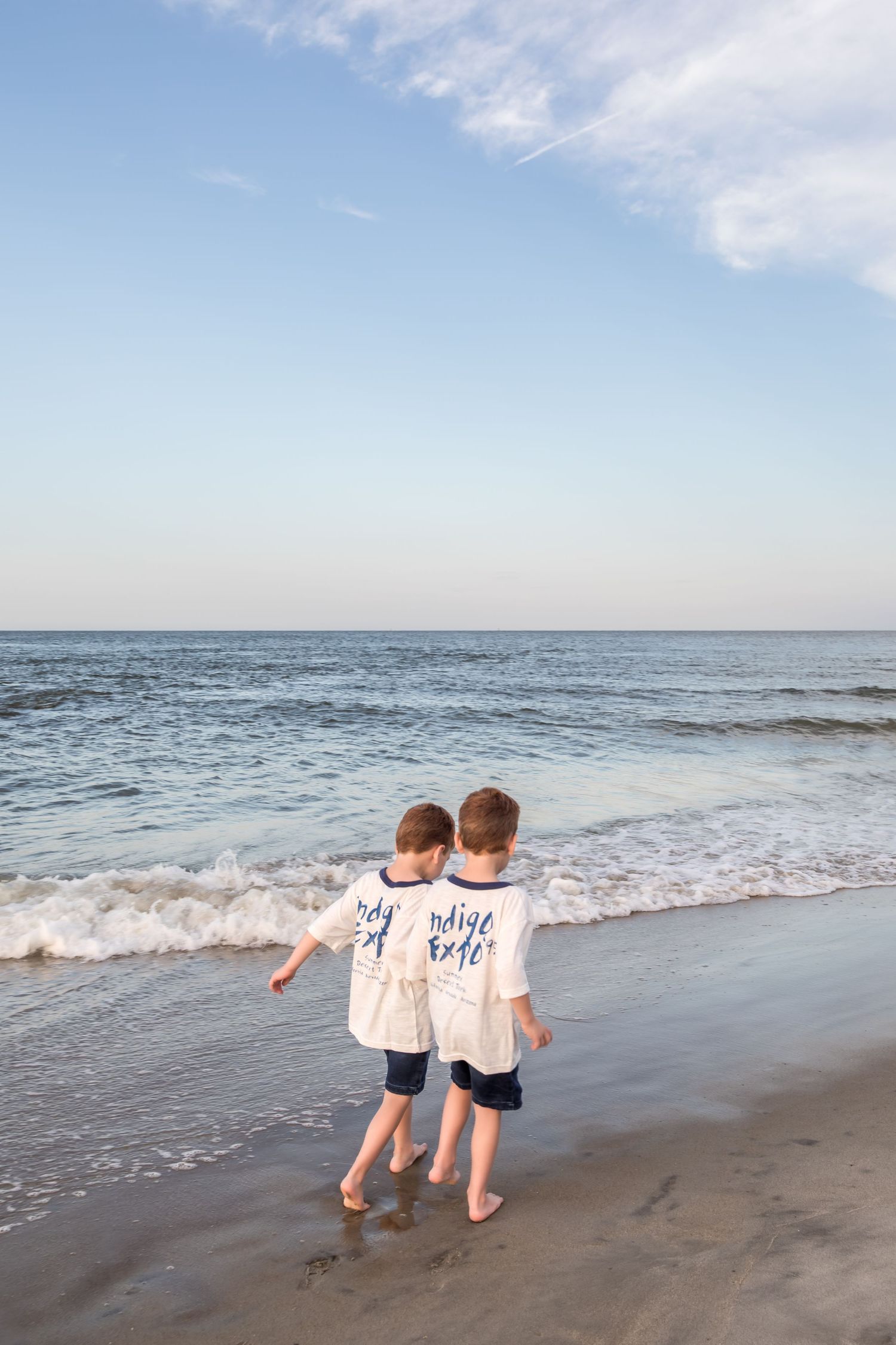 Two young boys in matching white shirts walk barefoot along a sandy ocean shoreline on a clear day.
