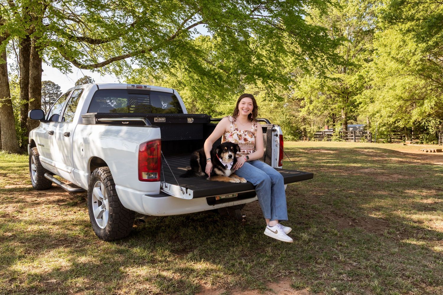 Woman sitting in truck bed with dog on green lawn under large tree.