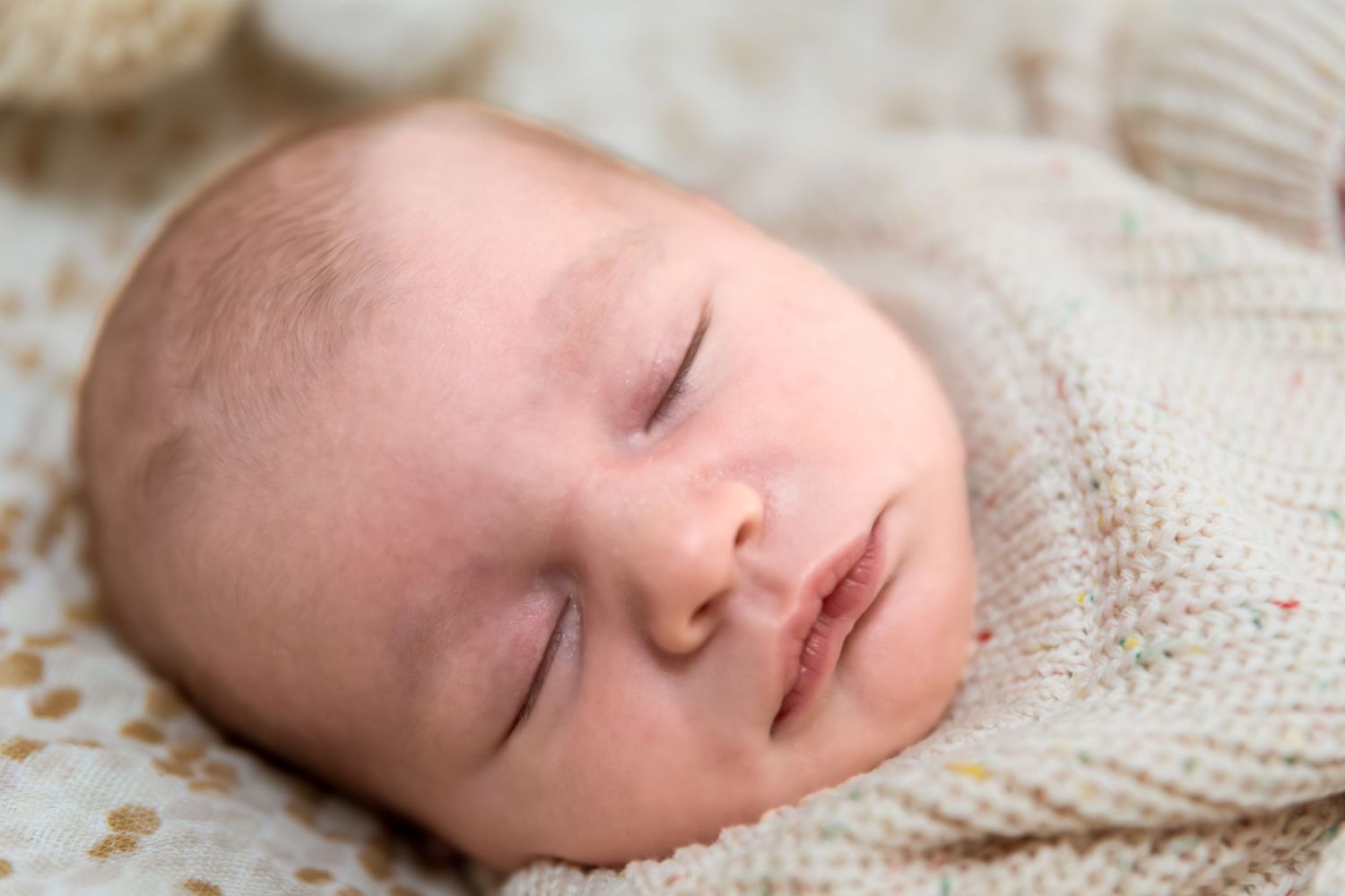 Peaceful newborn baby sleeping soundly, wrapped in a soft cream knit blanket with colorful speckles.