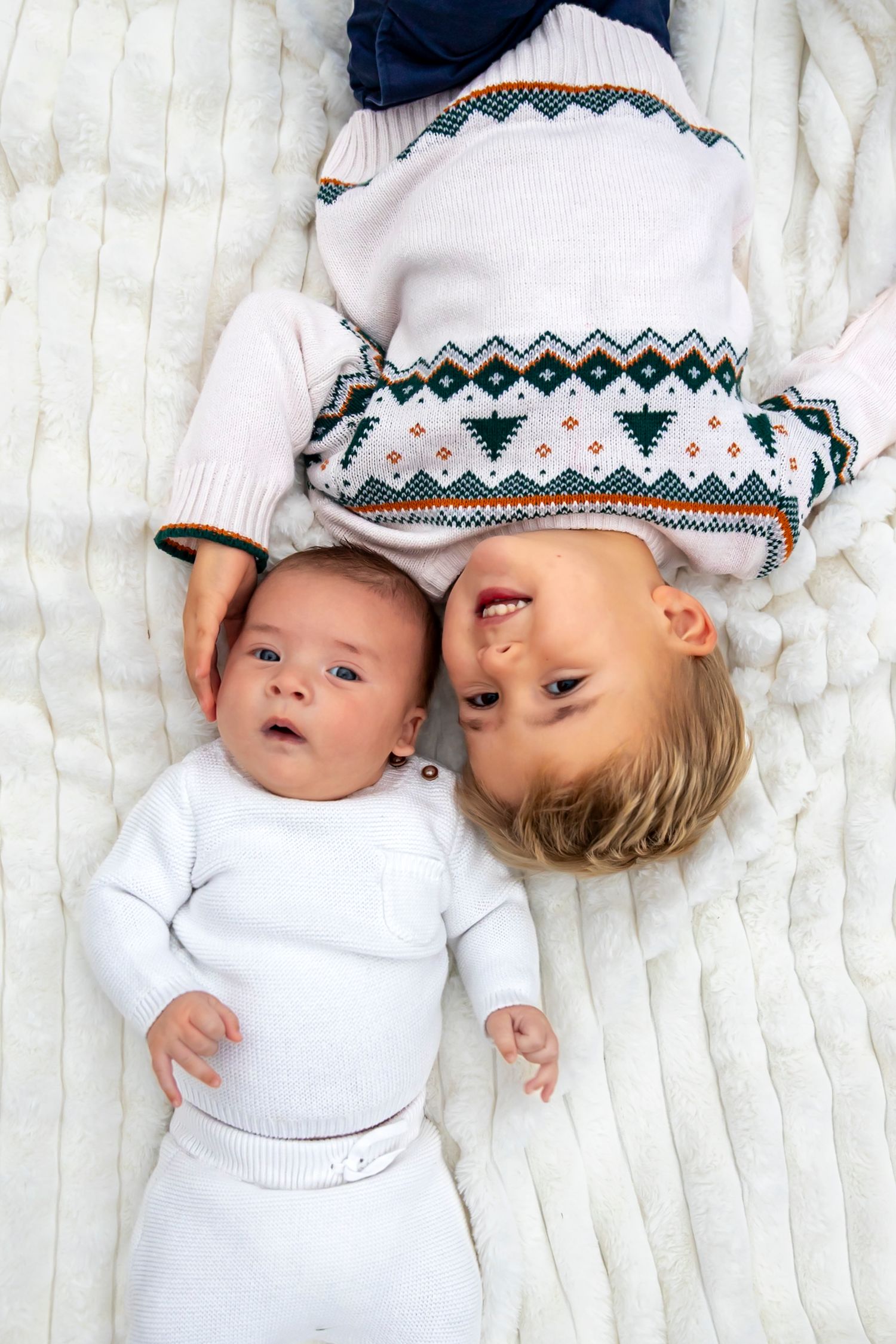 Two babies wearing matching winter sweaters lying on white blanket.