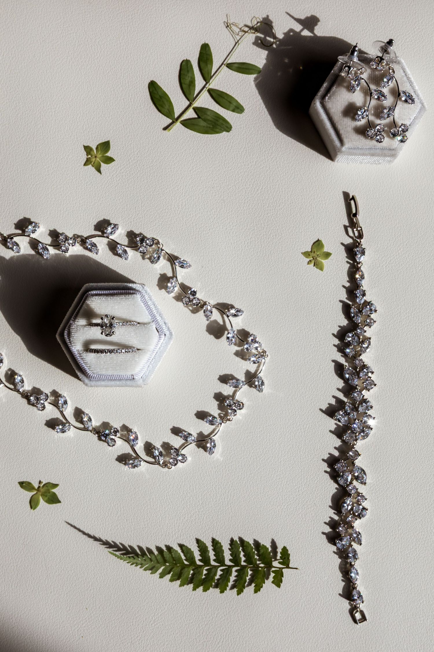 Wedding jewelry and green fern leaves arranged artistically on white background with hexagonal ring boxes and crystal bracelet.