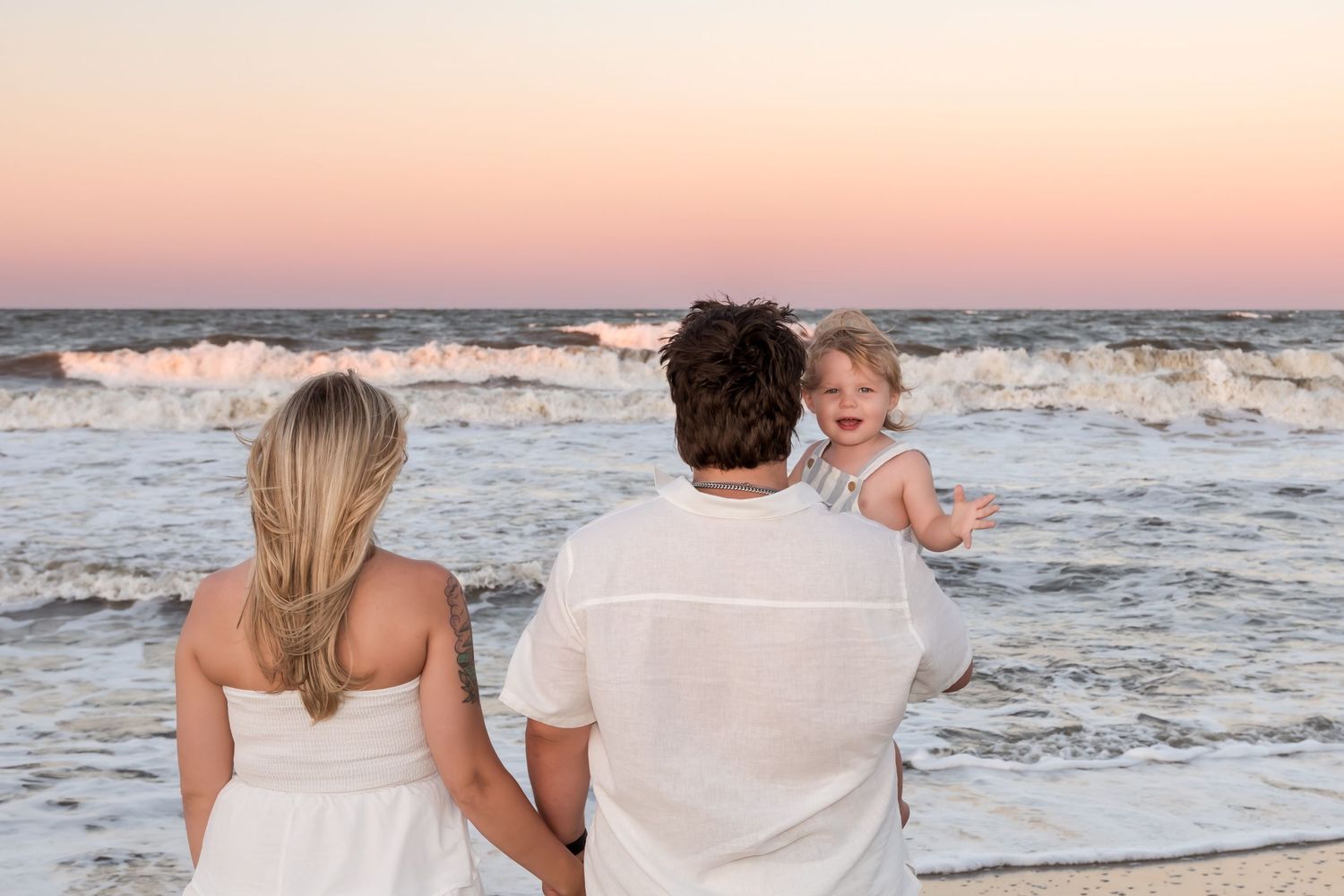 Family of three enjoying a sunset beach walk, parents holding toddler girl as waves crash on sandy shore.